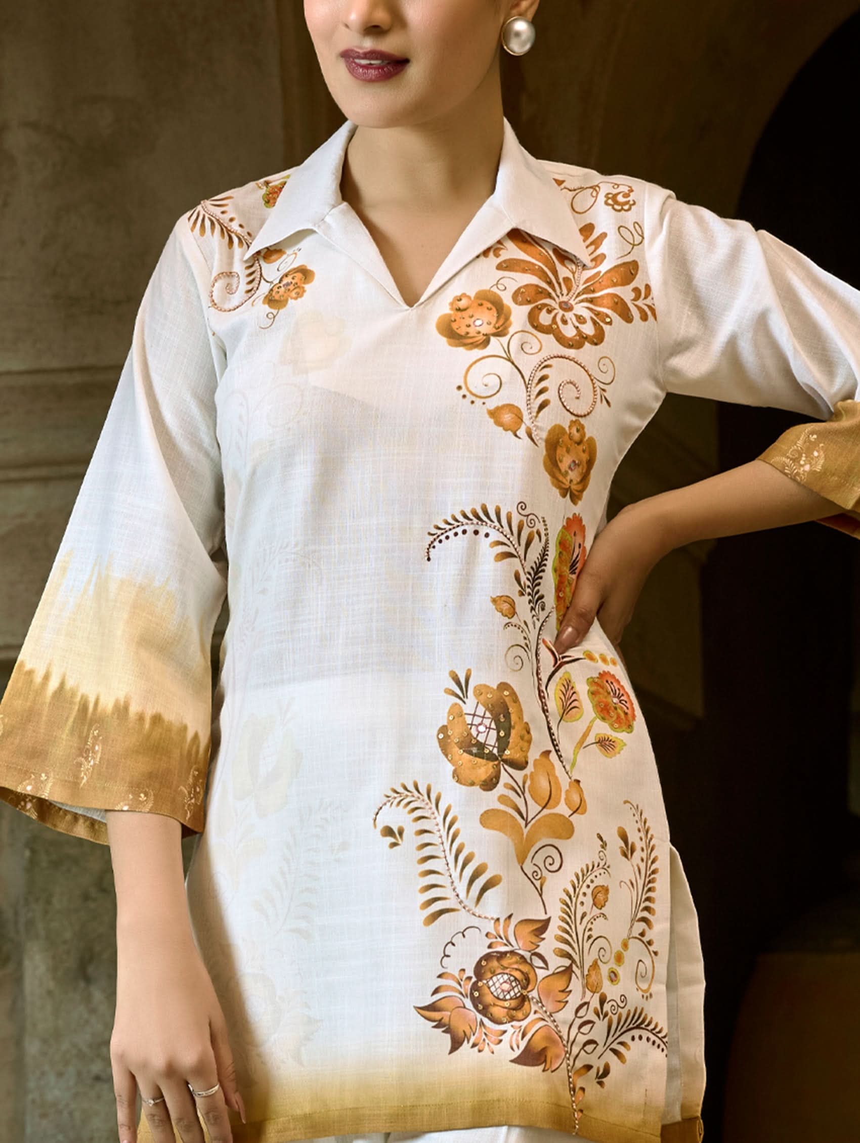 A woman wearing a digitally printed cotton linen short kurta with a revere collar and three-quarter sleeves, paired with straight pants. The outfit features white and golden colors with hand embroidery on the kurta.