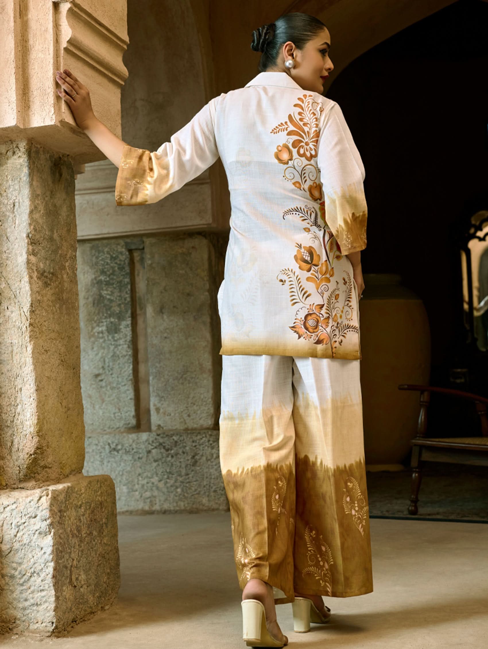 A woman wearing a digitally printed cotton linen short kurta with a revere collar and three-quarter sleeves, paired with straight pants. The outfit features white and golden colors with hand embroidery on the kurta.