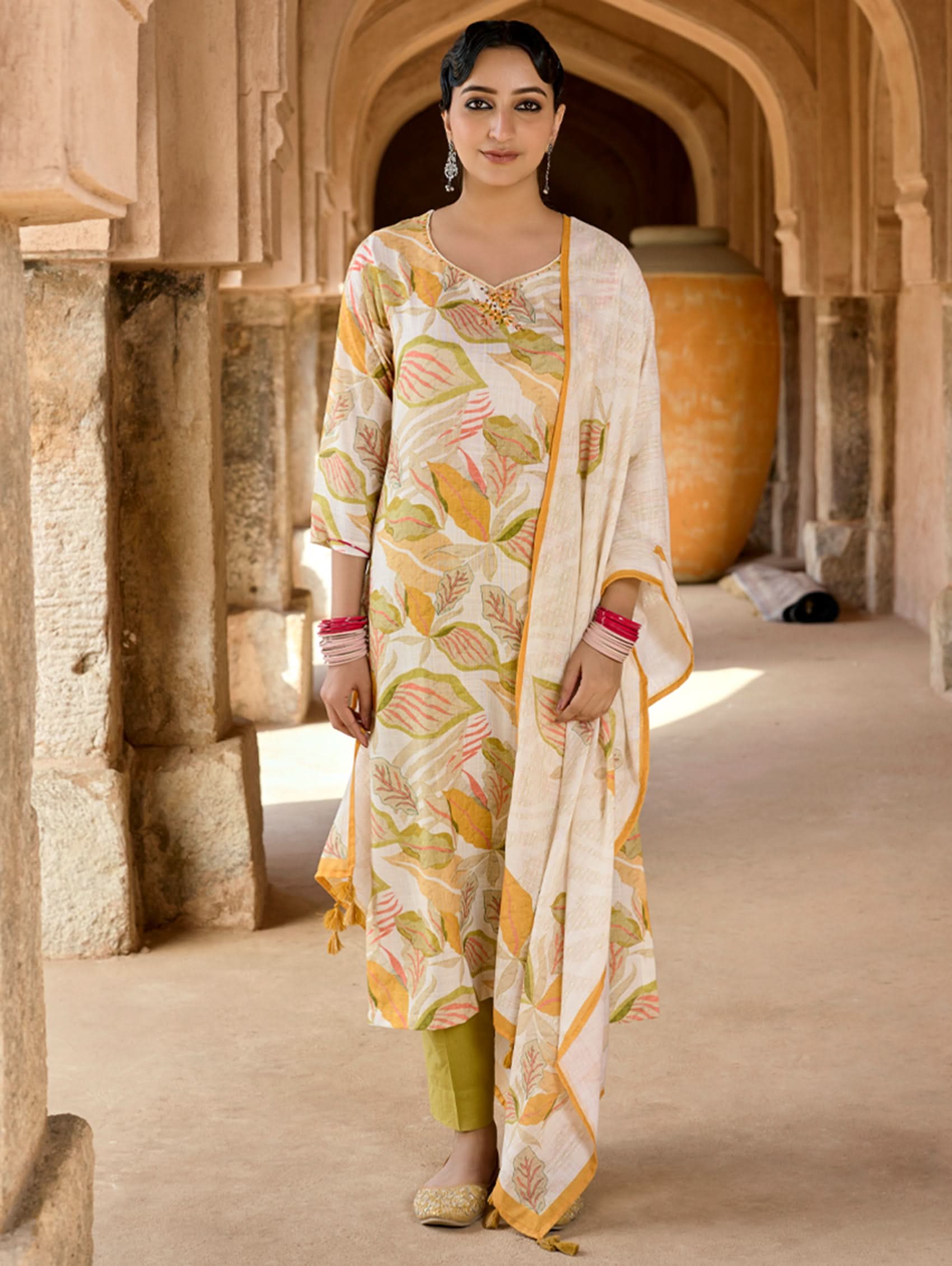 A woman standing in an archway wearing a digitally printed cotton linen suit set with a kurta, pants, and dupatta in beige, yellow, and green colors.