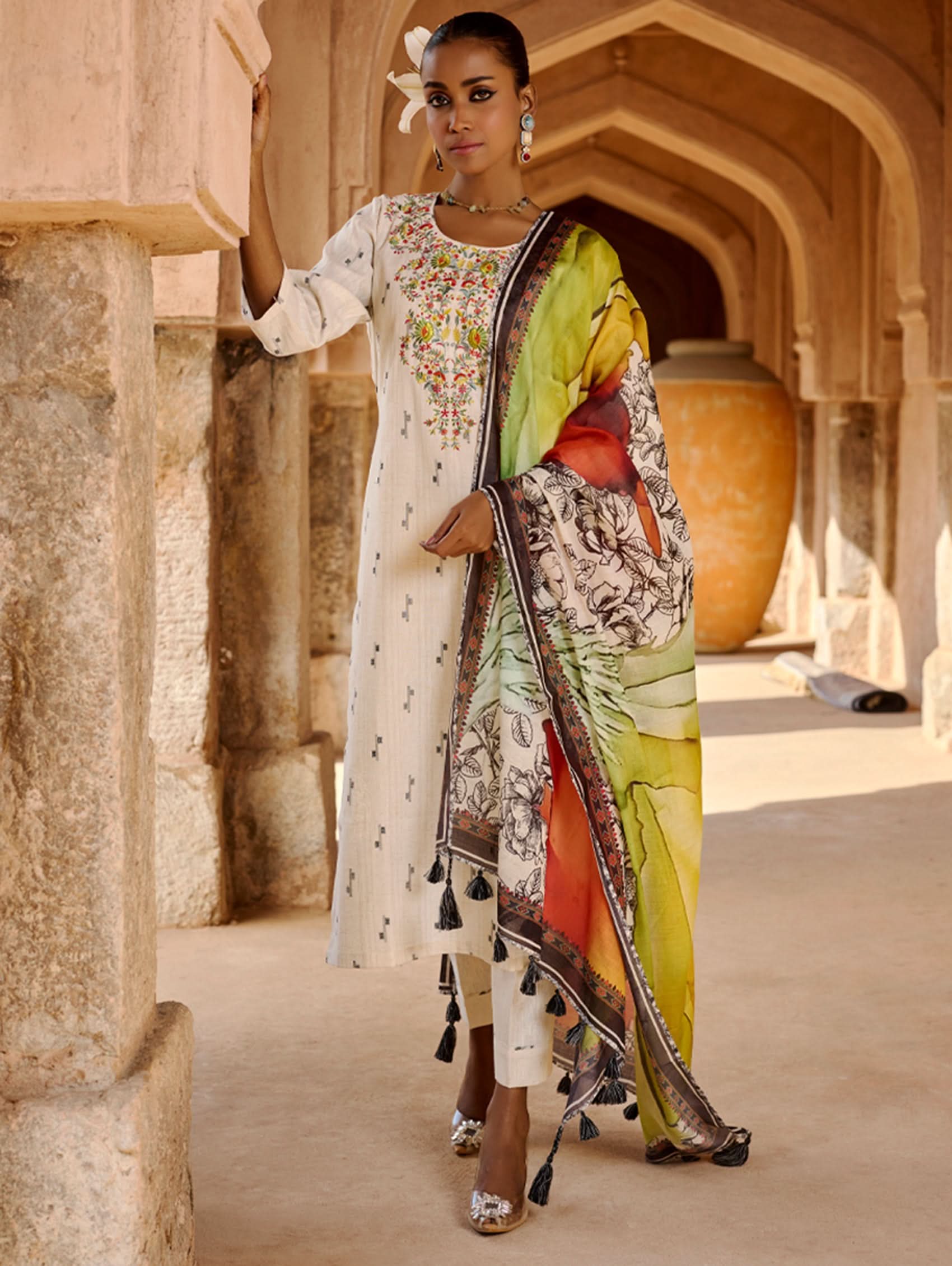 A woman modeling a white kurta with three-fourth sleeves, round neckline, and side slits, paired with straight pants and a multicolored dupatta with geometric patterns.