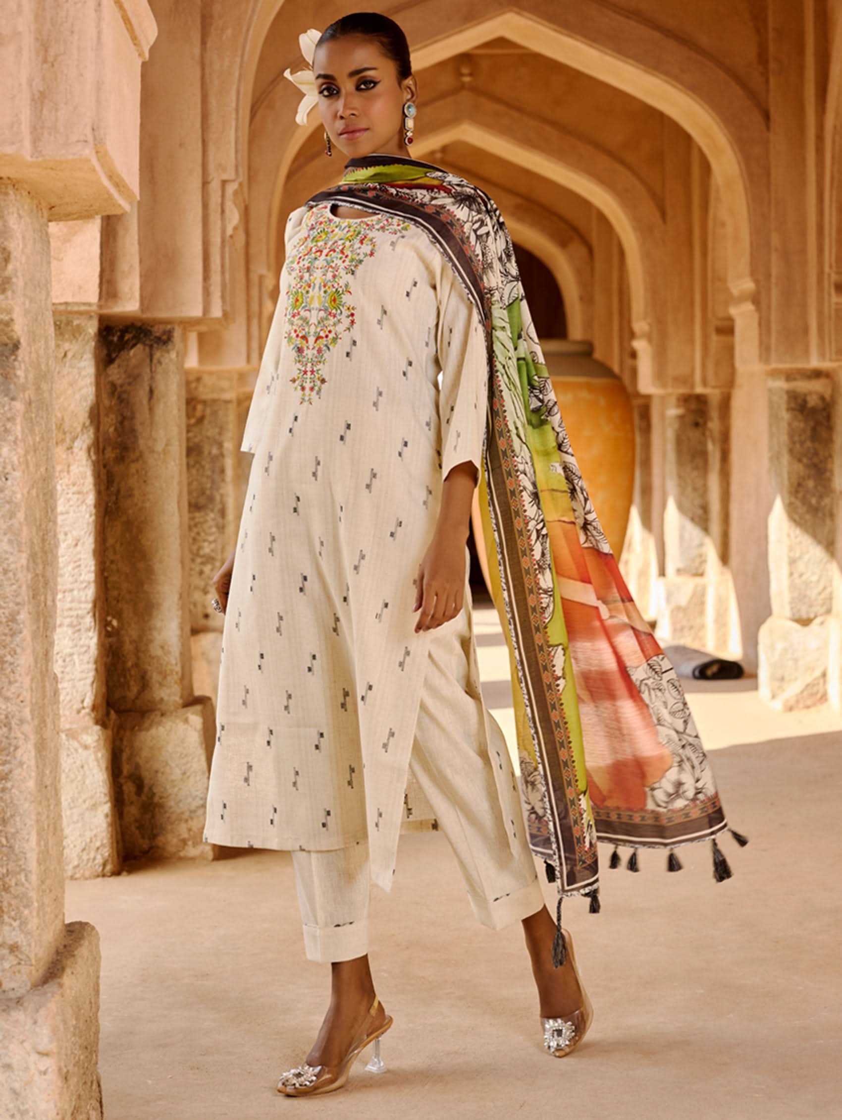 A woman modeling a white kurta with three-fourth sleeves, round neckline, and side slits, paired with straight pants and a multicolored dupatta with geometric patterns.