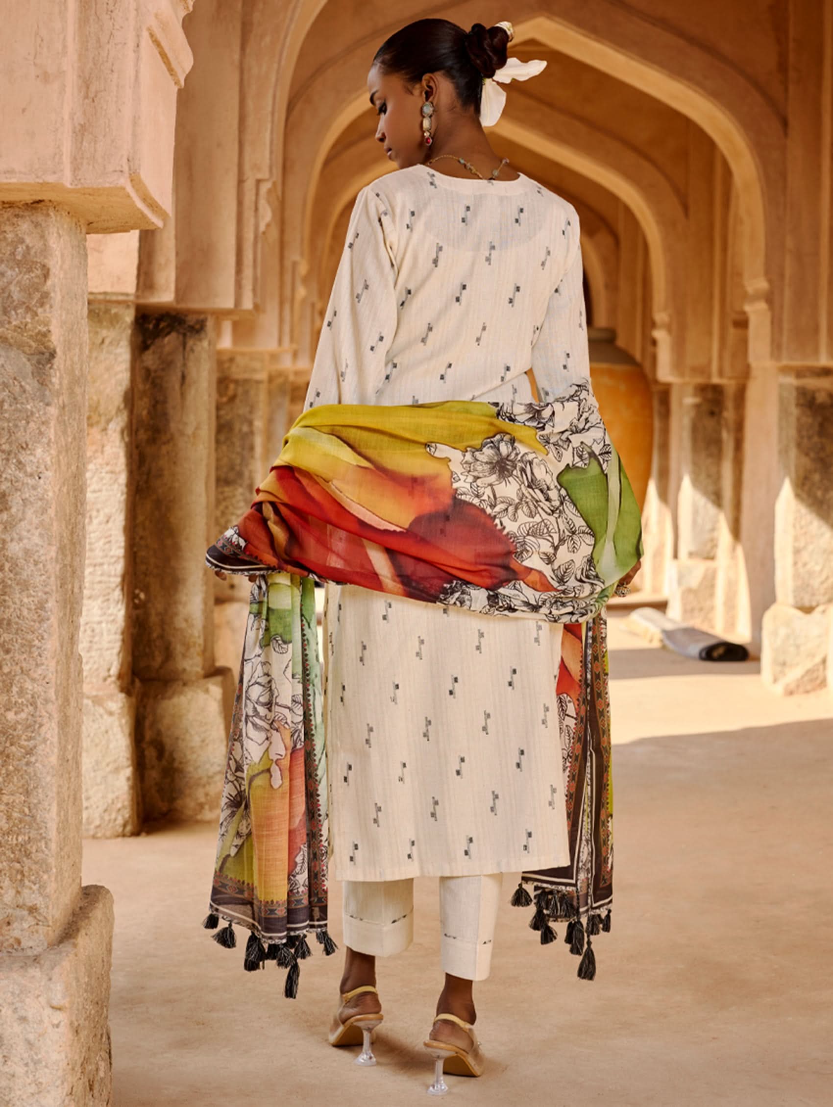 A woman modeling a white kurta with three-fourth sleeves, round neckline, and side slits, paired with straight pants and a multicolored dupatta with geometric patterns.