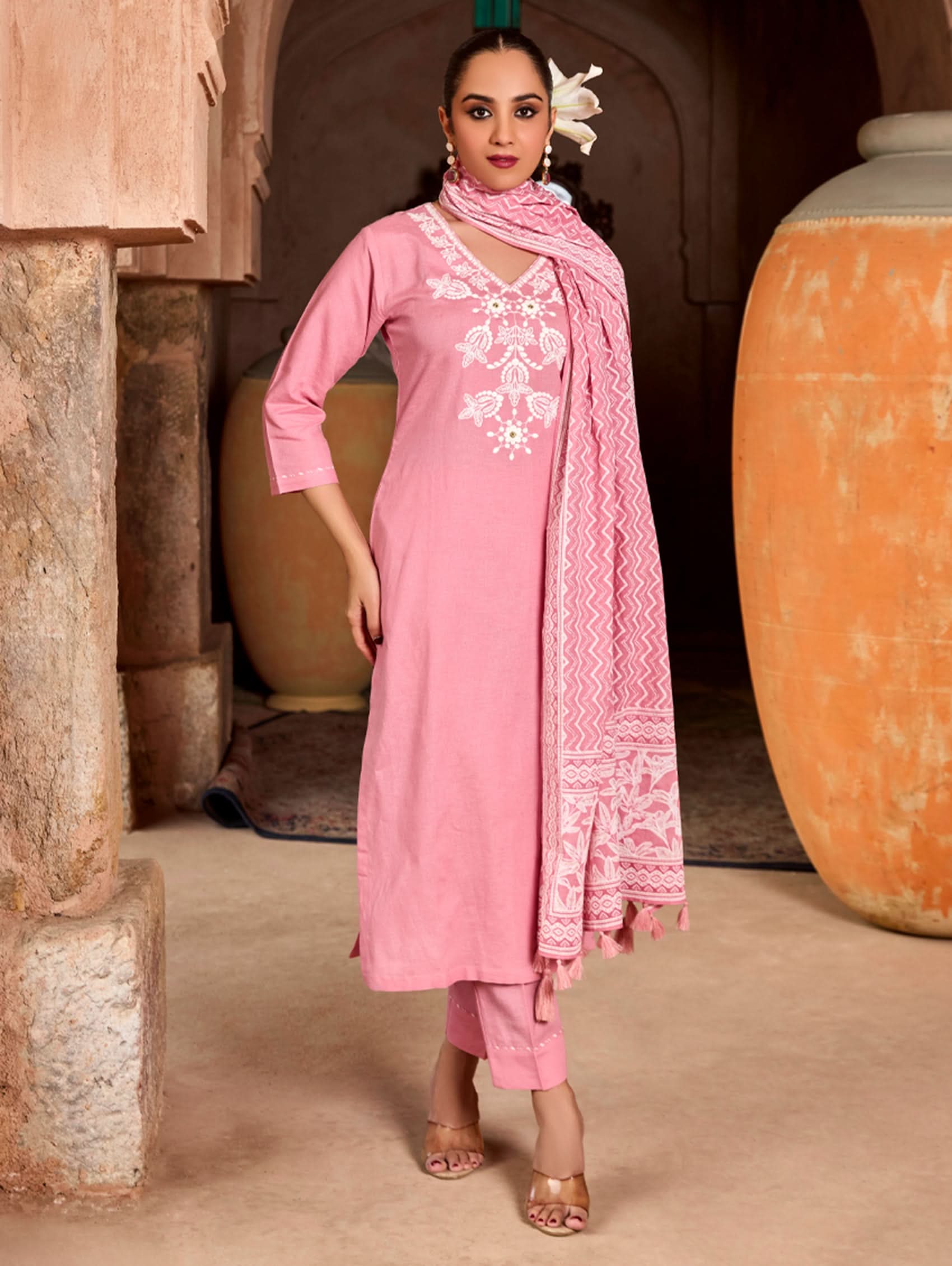 A woman wearing a pink suit set with embroidered details and a printed dupatta, standing against an rustic background.