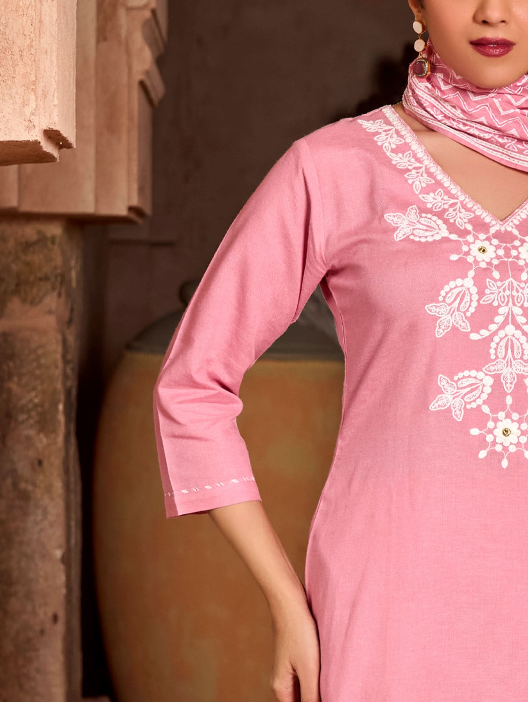 A woman wearing a pink suit set with embroidered details and a printed dupatta, standing against an rustic background.