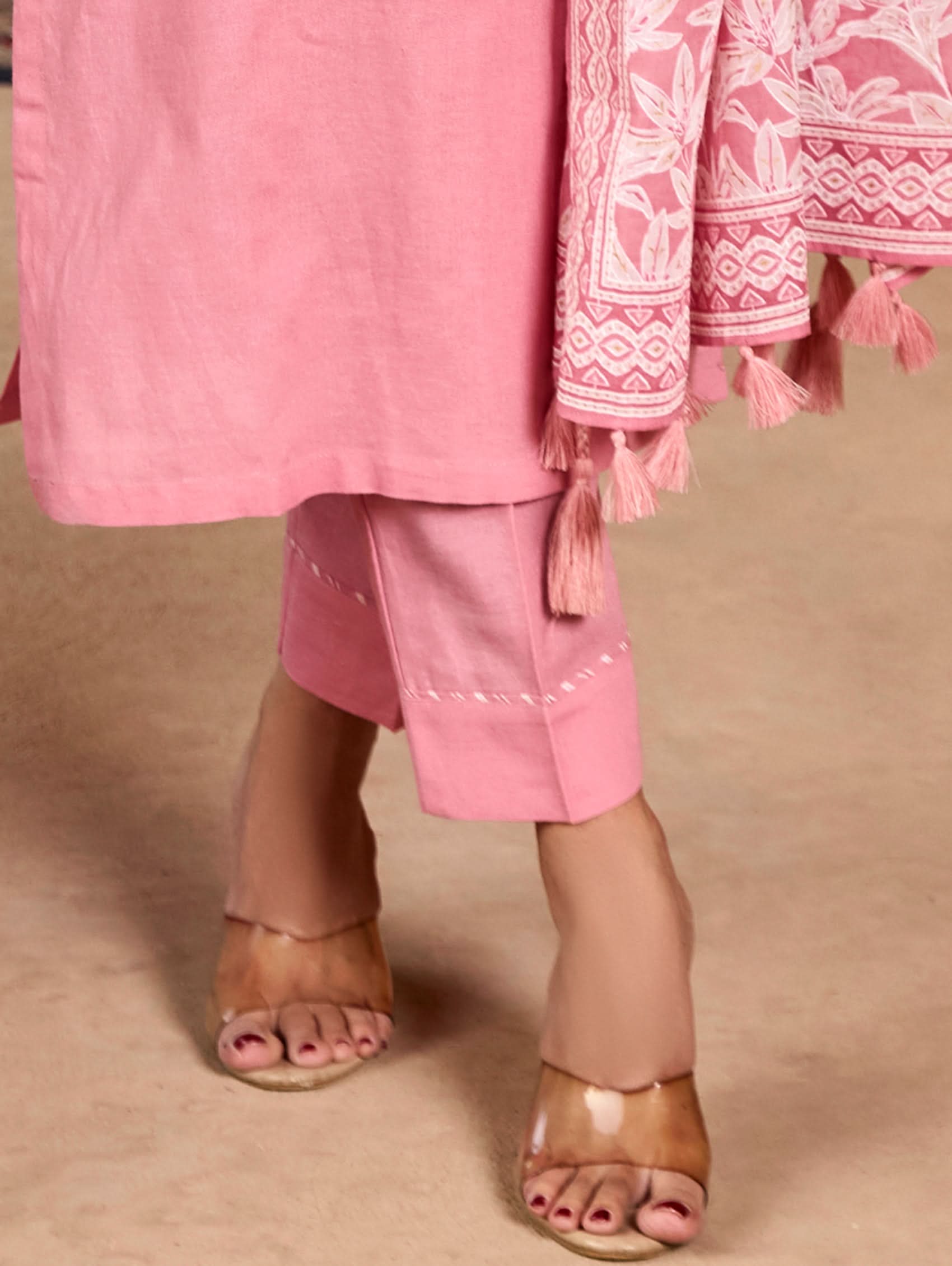 A woman wearing a pink suit set with embroidered details and a printed dupatta, standing against an rustic background.