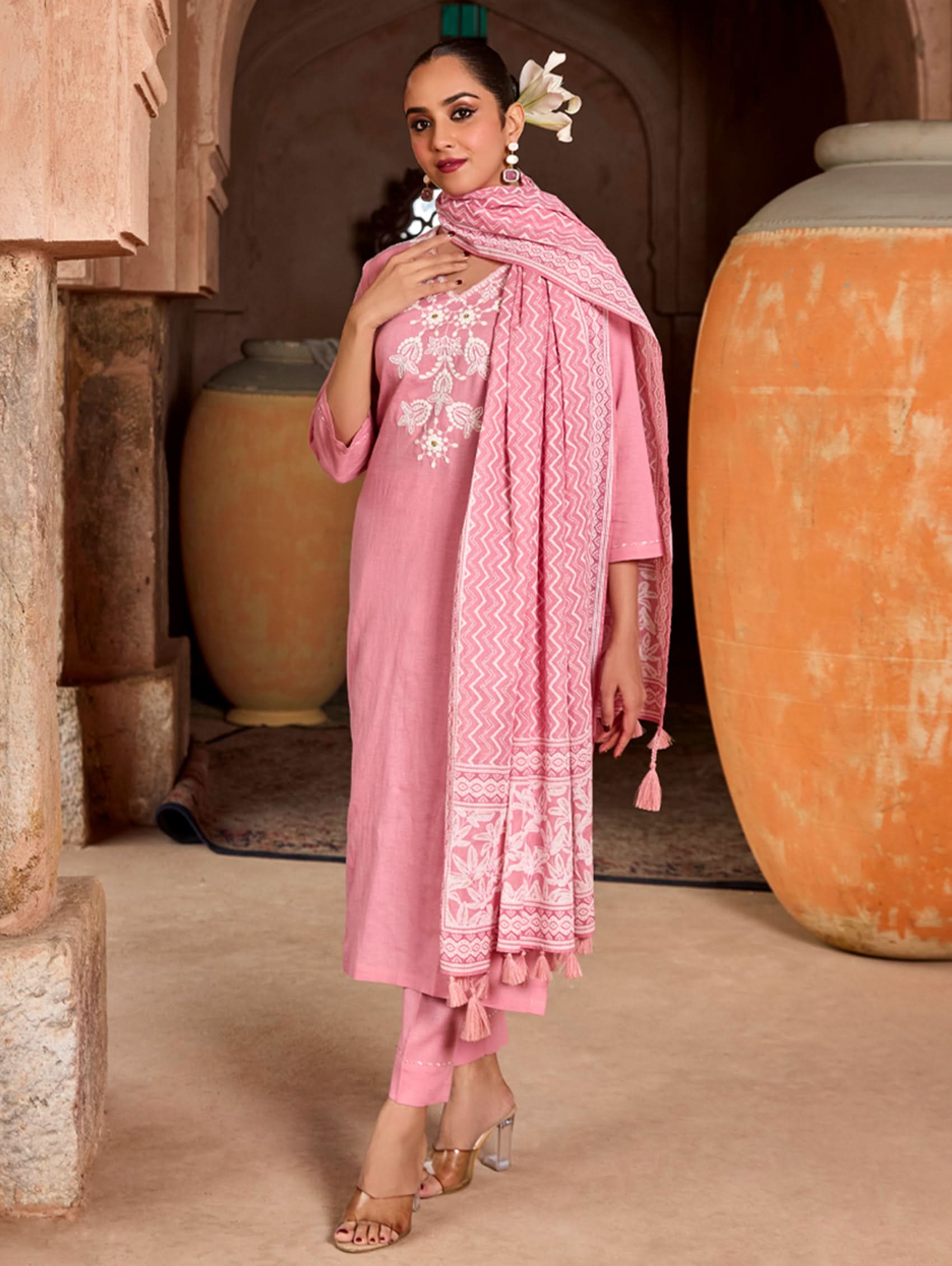 A woman wearing a pink suit set with embroidered details and a printed dupatta, standing against an rustic background.
