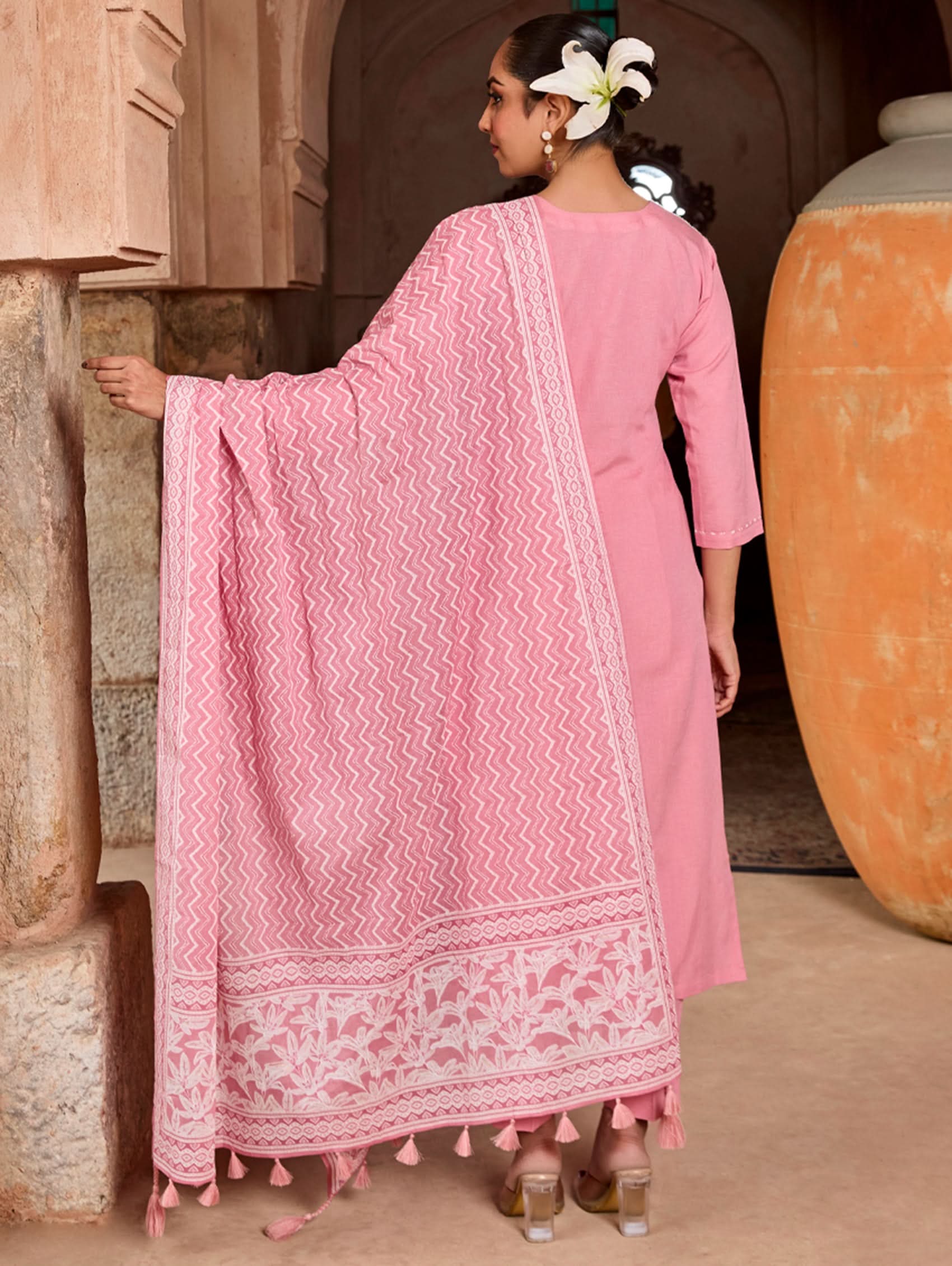 A woman wearing a pink suit set with embroidered details and a printed dupatta, standing against an rustic background.