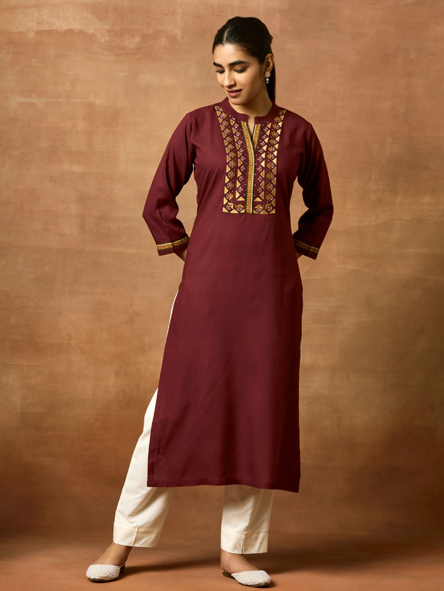 A woman plus-size model wearing a maroon viscose rayon kurta with a ban collar, three-fourth sleeves, and side slits. The kurta has intricate computerised thread embroidery on the chest area.