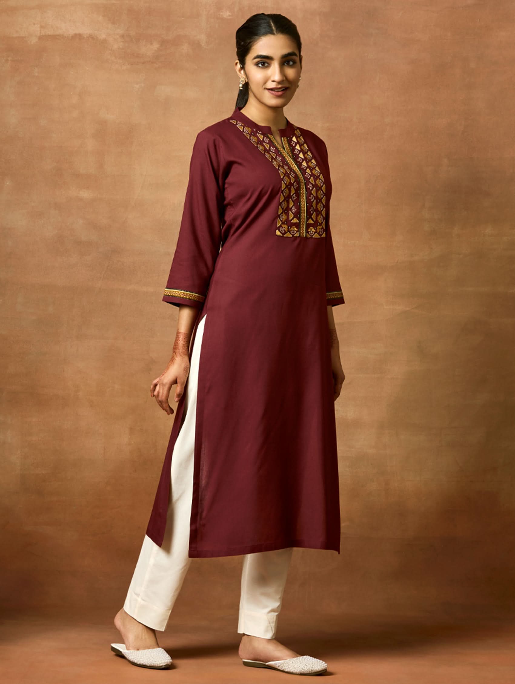 A woman plus-size model wearing a maroon viscose rayon kurta with a ban collar, three-fourth sleeves, and side slits. The kurta has intricate computerised thread embroidery on the chest area.