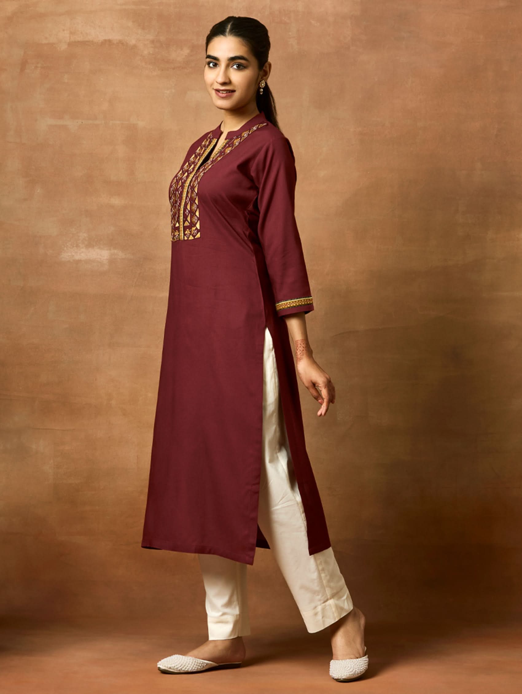 A woman plus-size model wearing a maroon viscose rayon kurta with a ban collar, three-fourth sleeves, and side slits. The kurta has intricate computerised thread embroidery on the chest area.