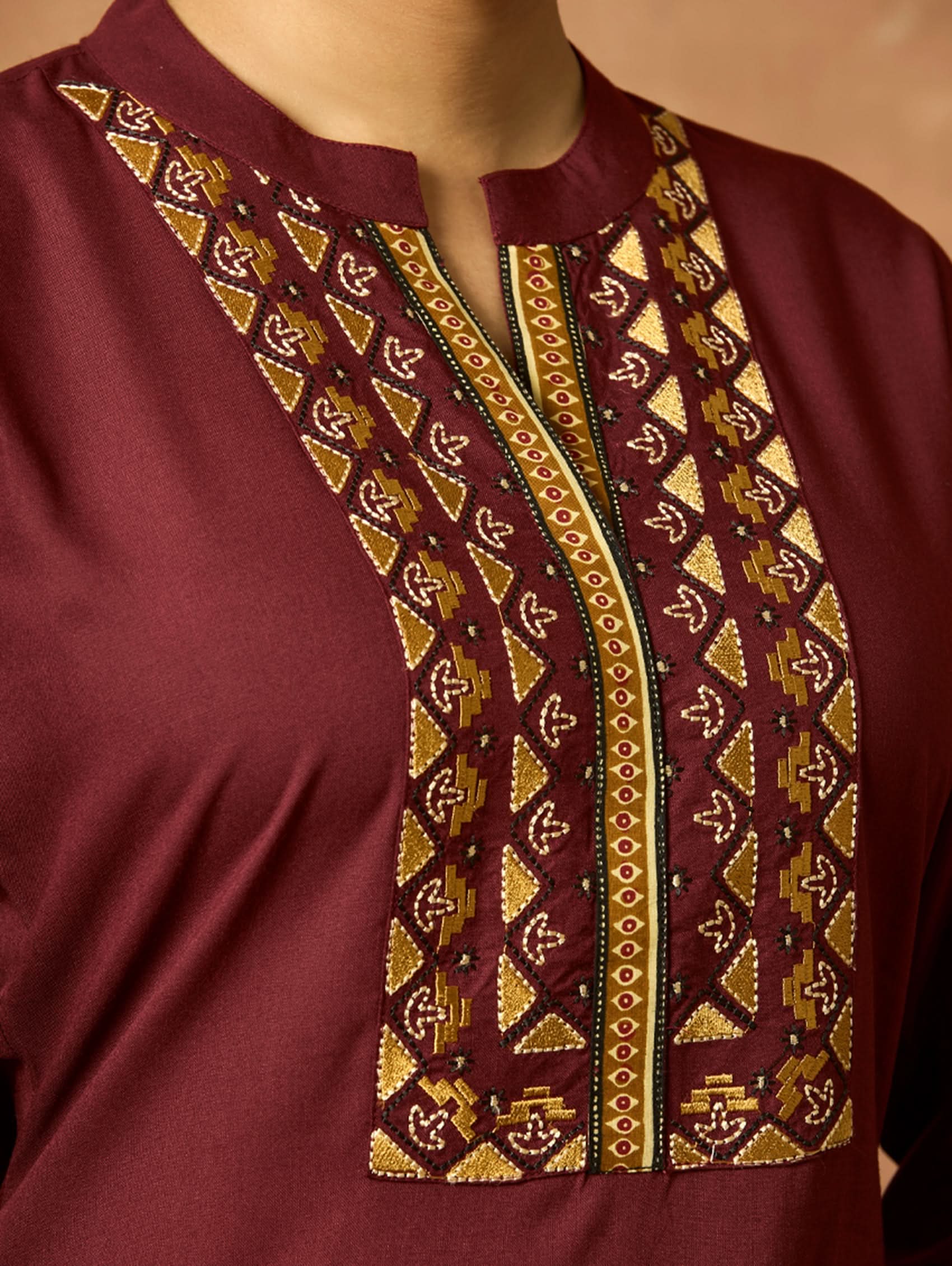 A woman plus-size model wearing a maroon viscose rayon kurta with a ban collar, three-fourth sleeves, and side slits. The kurta has intricate computerised thread embroidery on the chest area.