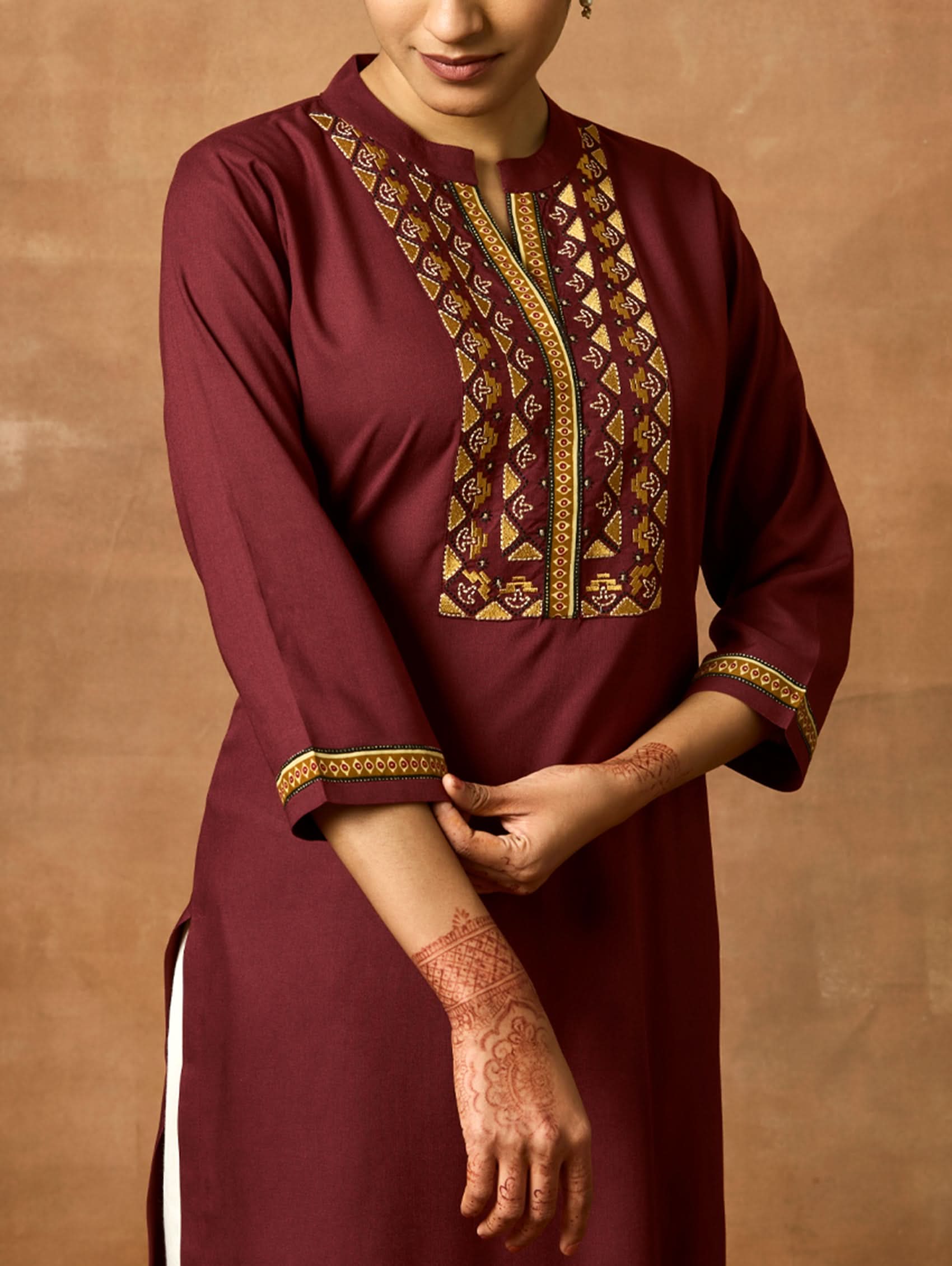 A woman plus-size model wearing a maroon viscose rayon kurta with a ban collar, three-fourth sleeves, and side slits. The kurta has intricate computerised thread embroidery on the chest area.