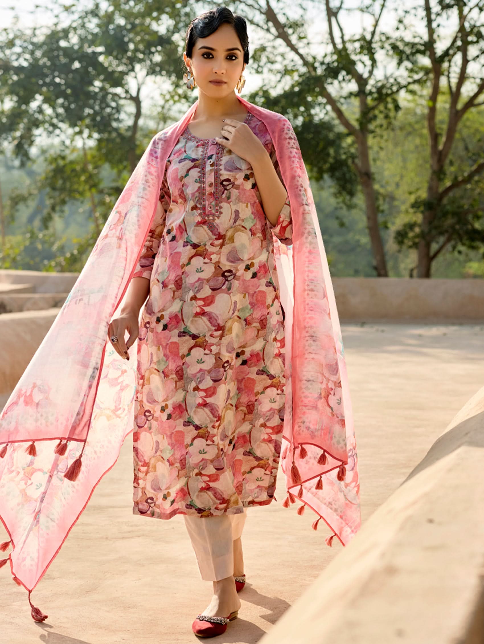 A woman standing in an archway, wearing a digitally printed cotton linen suit set with a round neckline kurta, three-fourth sleeves, side slits, and a dupatta with a floral pattern.