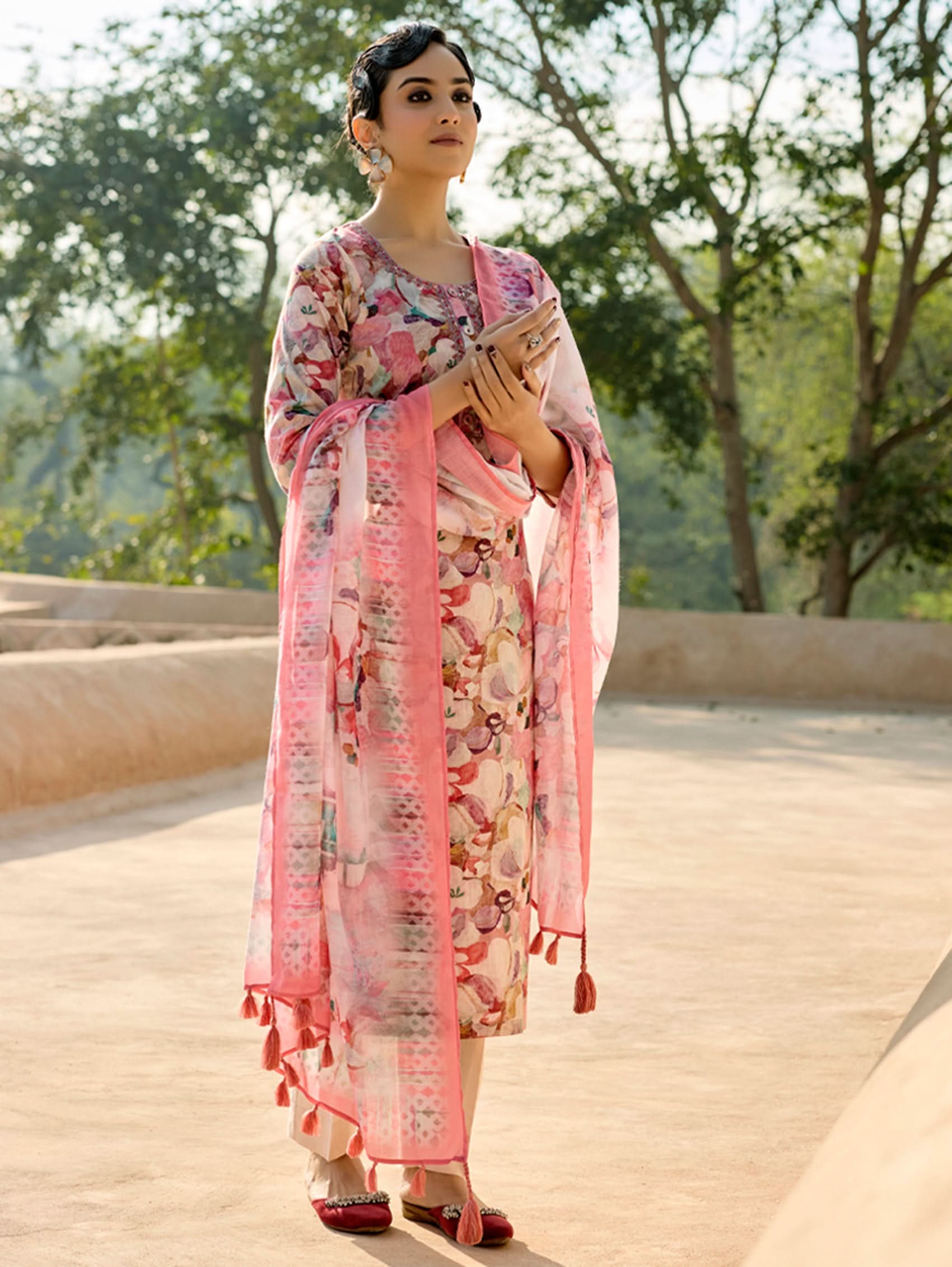 A woman standing in an archway, wearing a digitally printed cotton linen suit set with a round neckline kurta, three-fourth sleeves, side slits, and a dupatta with a floral pattern.