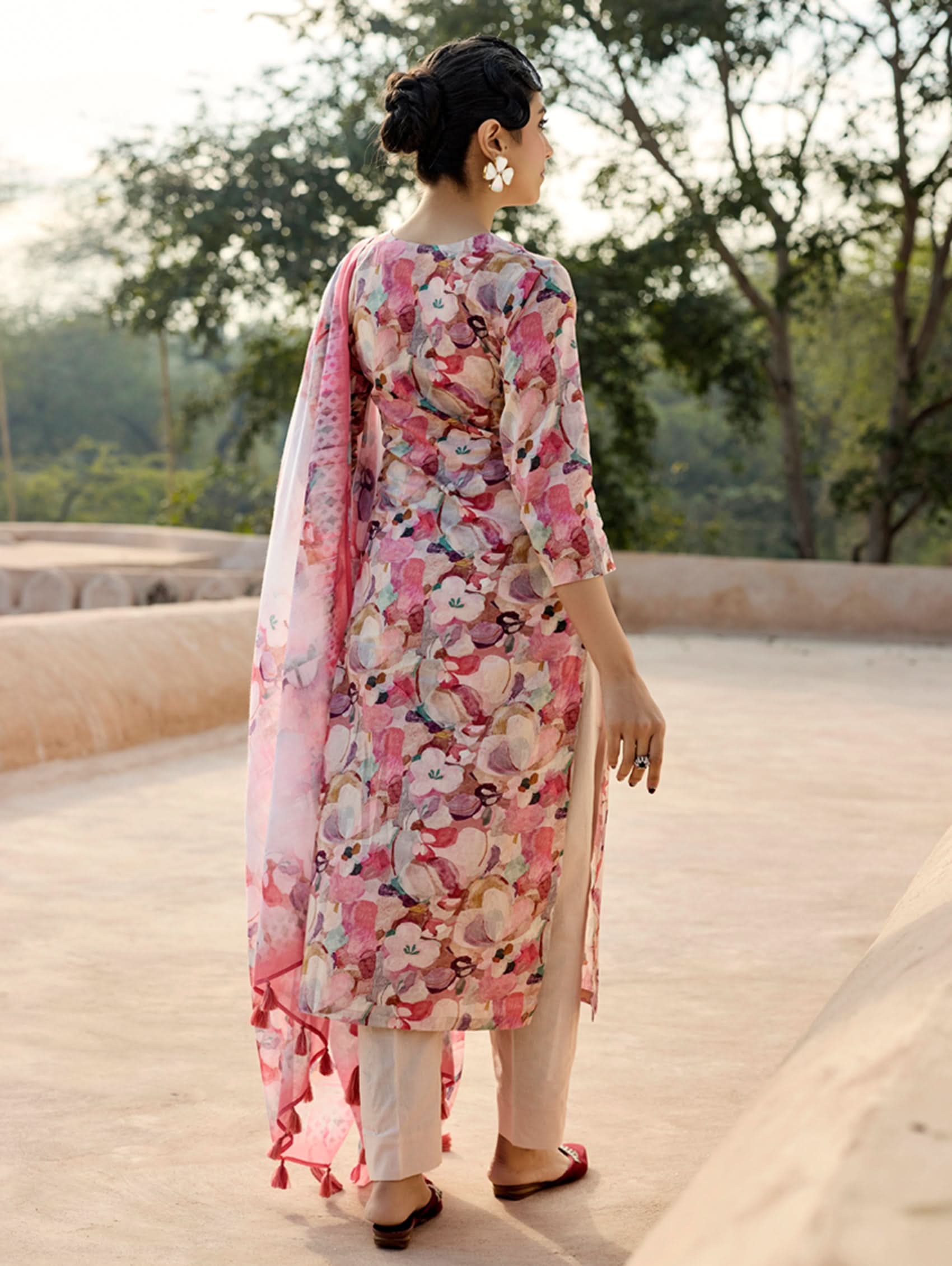 A woman standing in an archway, wearing a digitally printed cotton linen suit set with a round neckline kurta, three-fourth sleeves, side slits, and a dupatta with a floral pattern.
