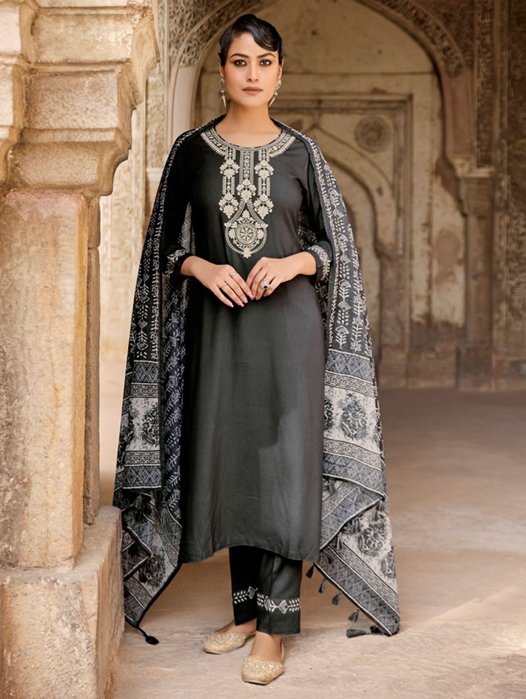 A woman standing in a historic location wearing a traditional grey suit set with embroidery and a printed dupatta.