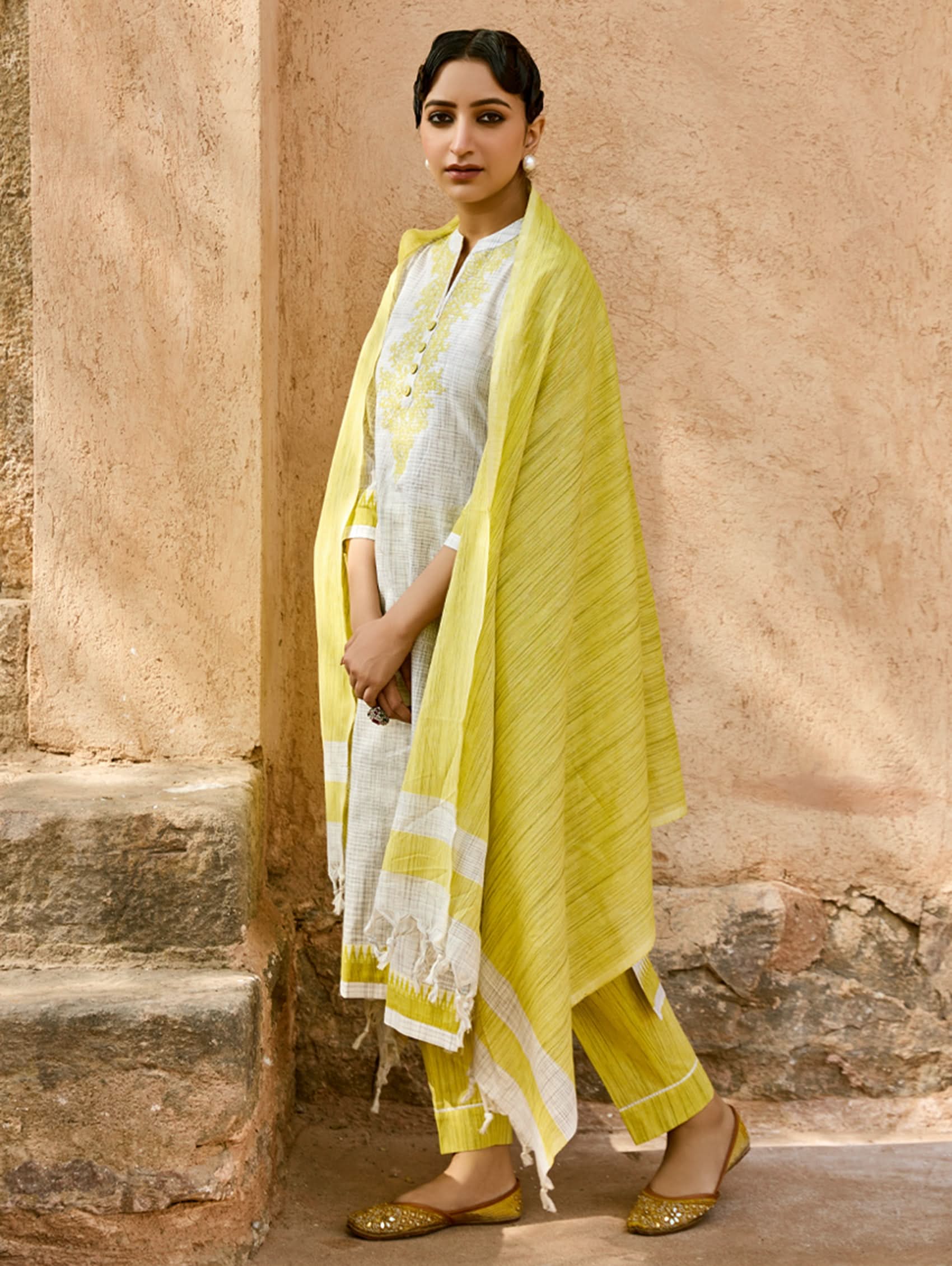 Woman in a yellow and white traditional outfit standing against a beige wall.