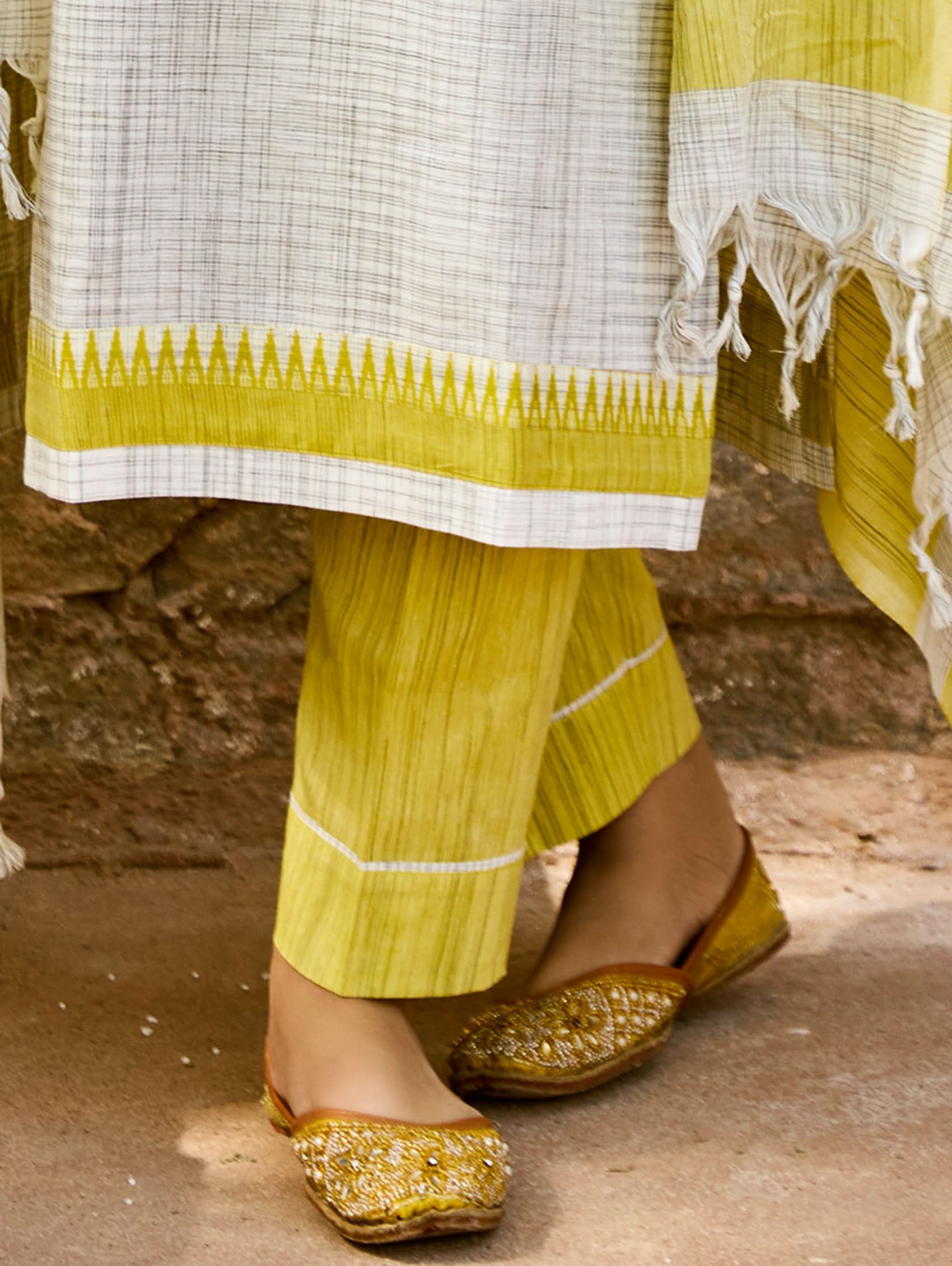 Traditional outfit with a white and yellow patterned top and pants, paired with gold sandals on a rustic background.