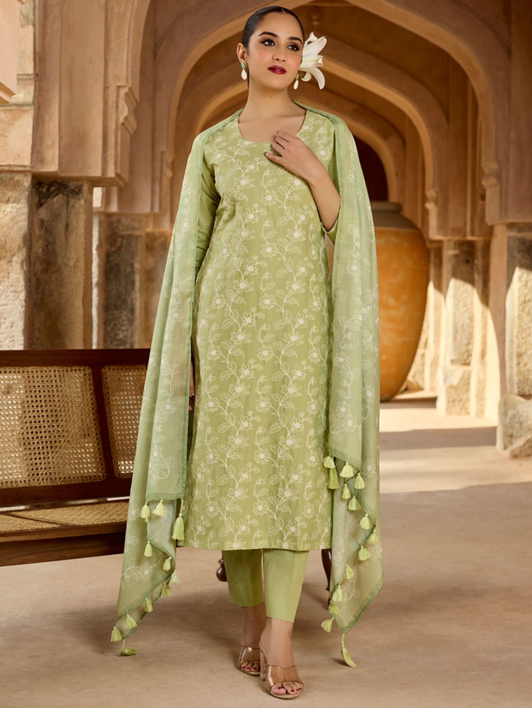 A woman wearing a green embroidered pure cotton suit set with a kurta, pants, and a dupatta, standing in a traditional setting.
