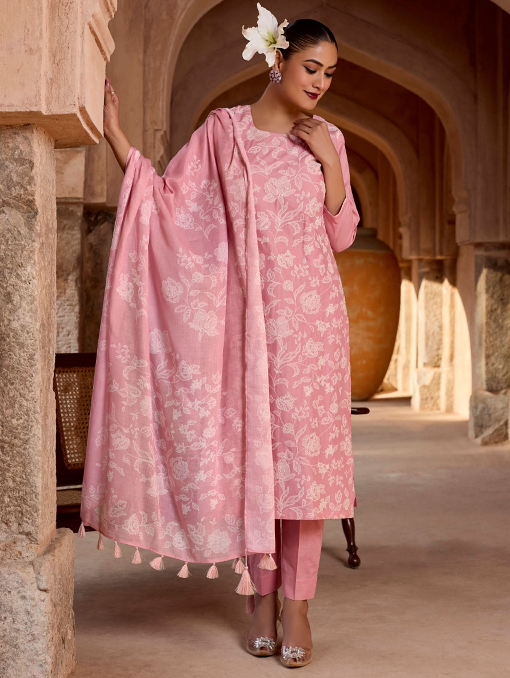 A woman standing in a pose wearing a pink floral embroidered pure cotton suit set with a kurta, pants, and a dupatta.