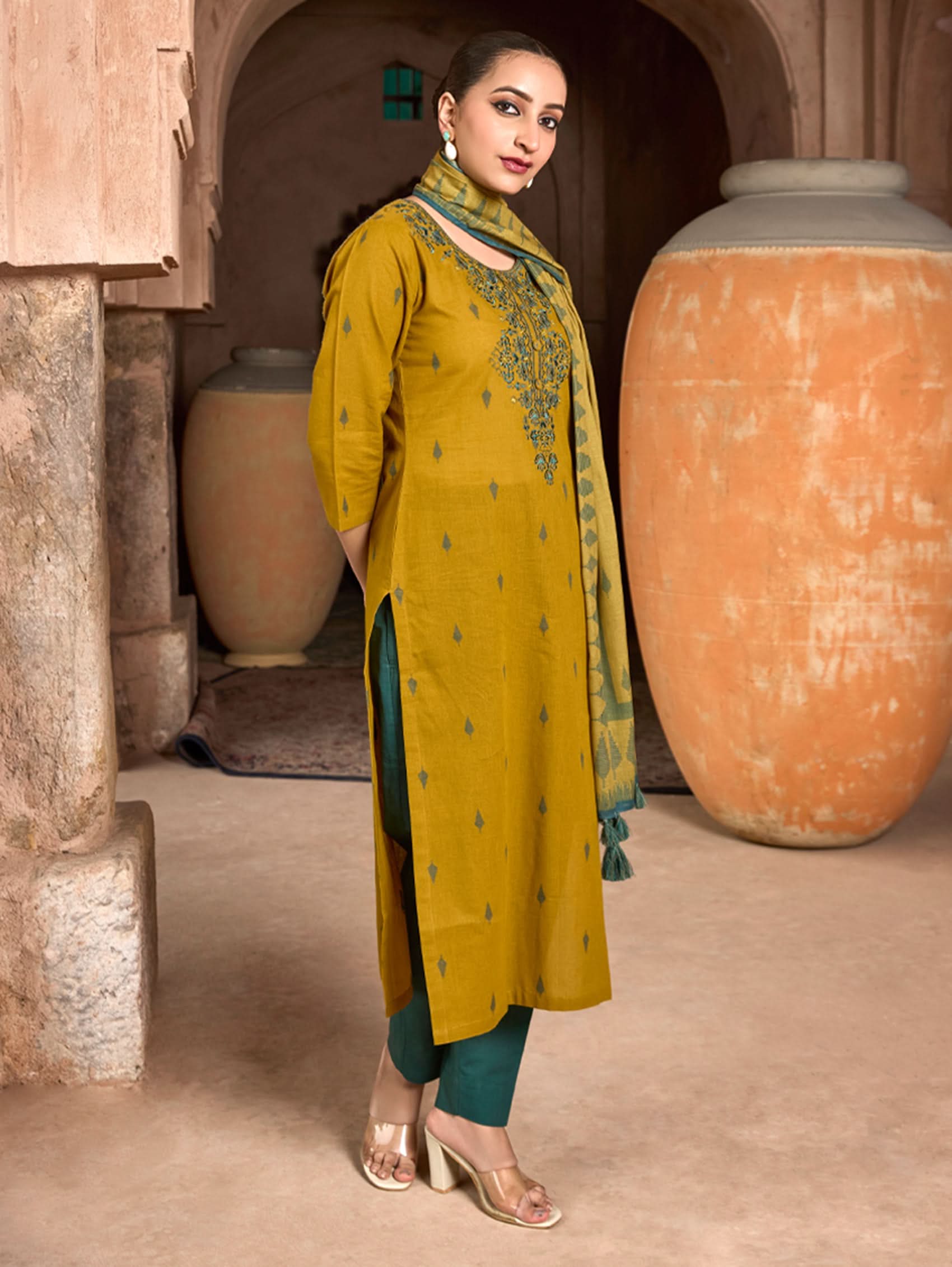 Woman in yellow kurta and green dupatta standing in a rustic setting.