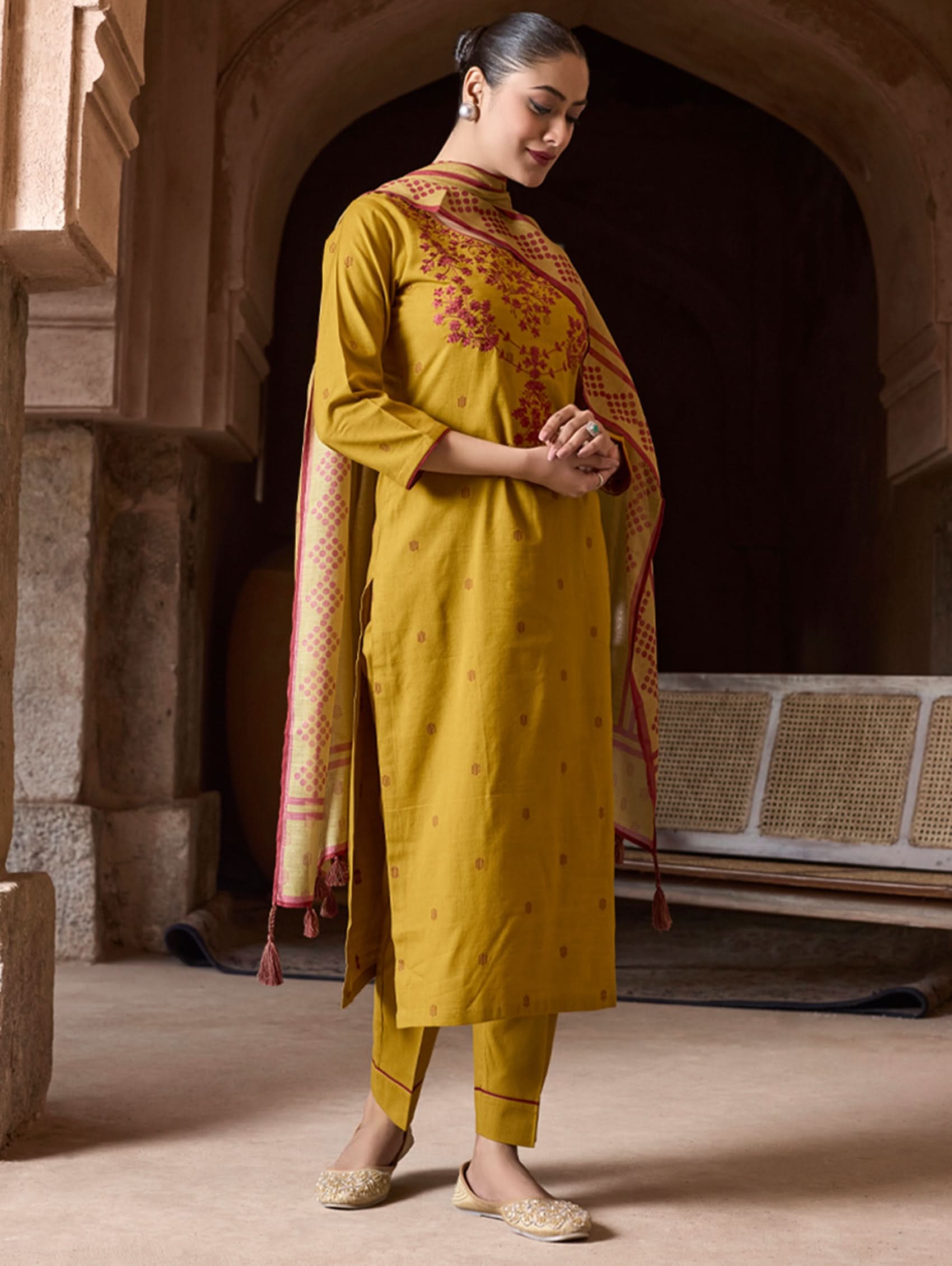 Woman in a mustard yellow traditional outfit with a red and gold dupatta standing in an archway.