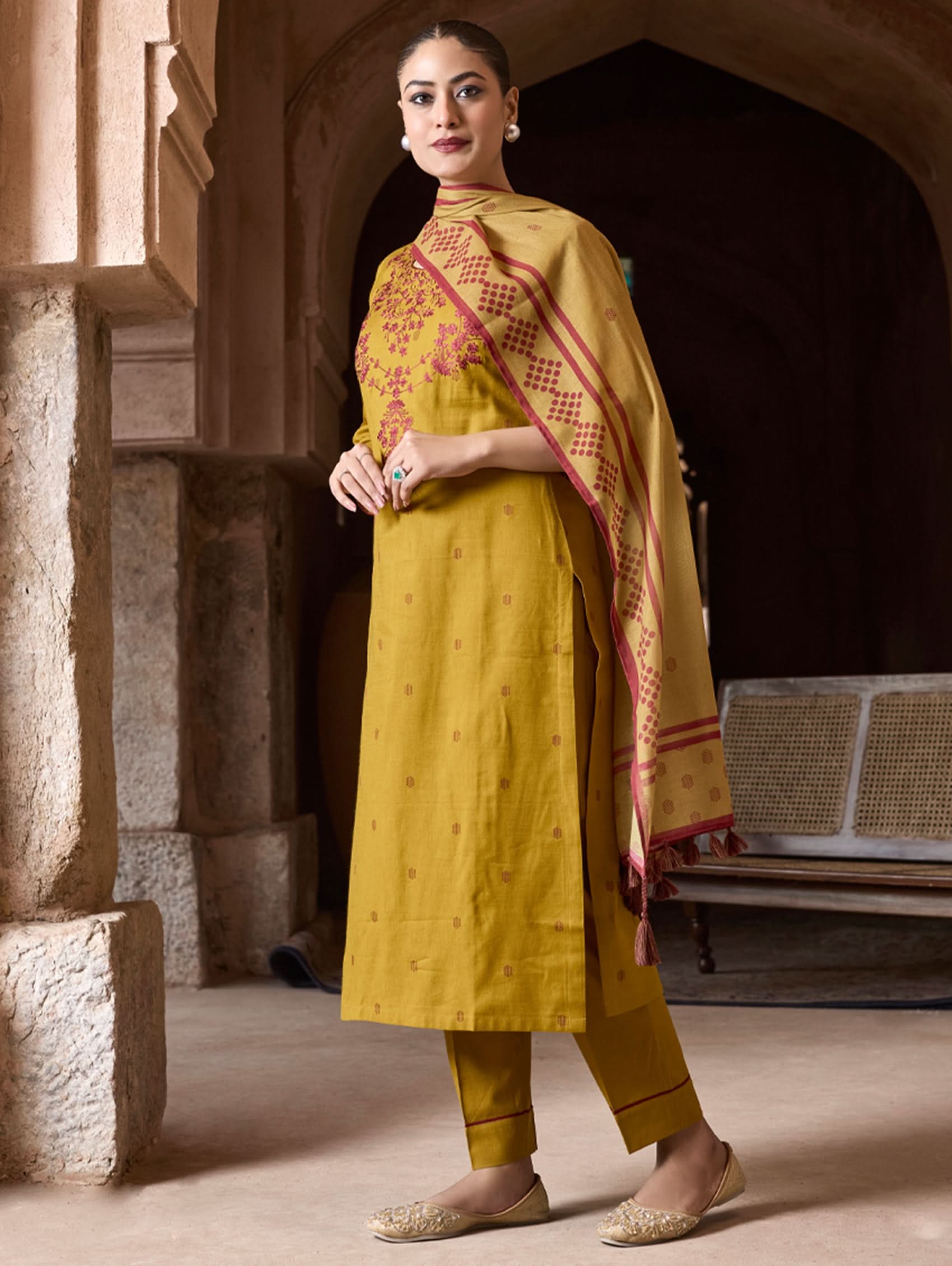 Woman in a yellow traditional outfit with a red and gold dupatta standing in an architectural setting.