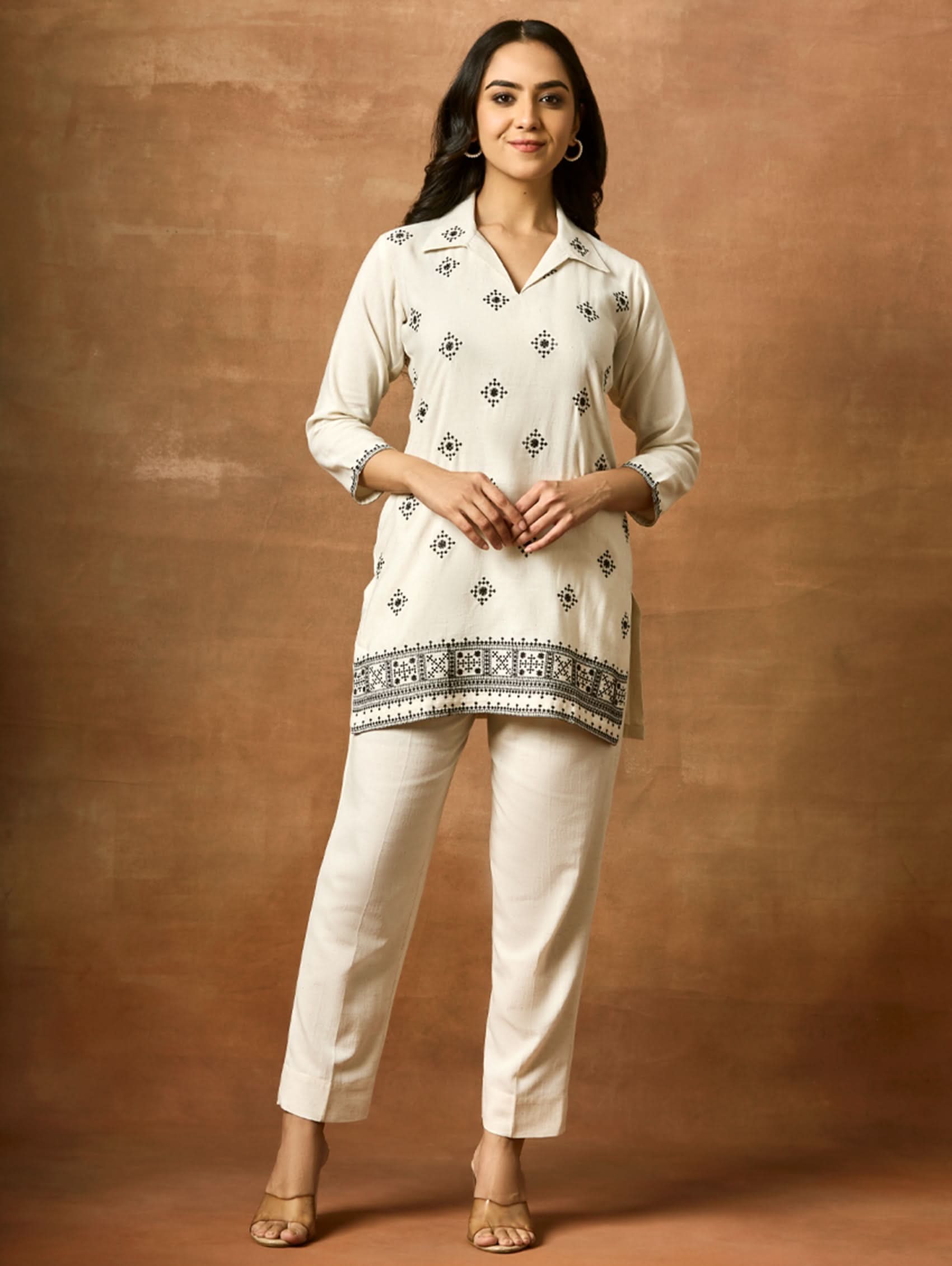 A woman modeling a white and blue embroidered kurta set with three-fourth sleeves and a revere collar, paired with regular pants, against a brown backdrop.