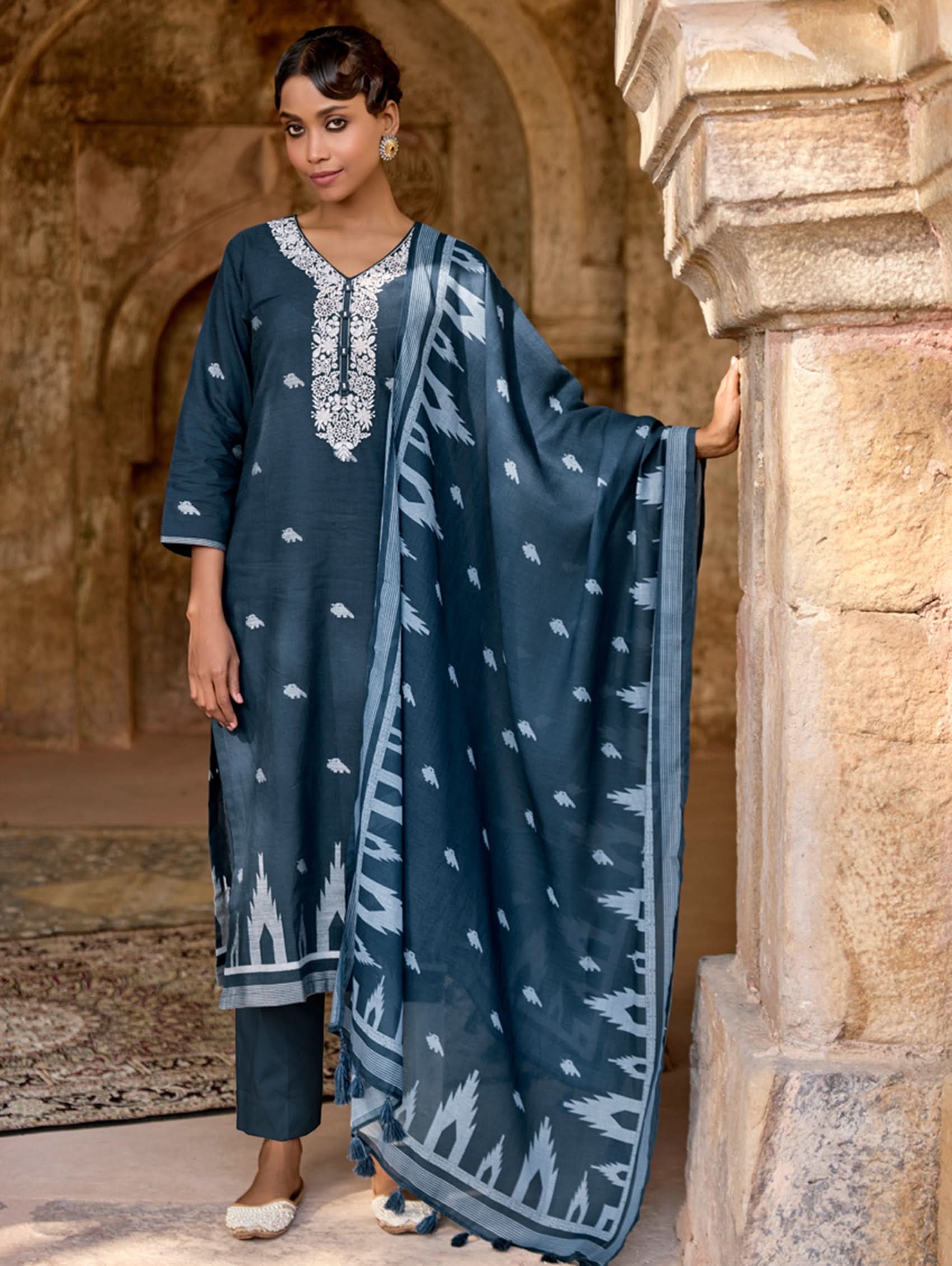A woman standing in an archway, wearing a blue and white floral print kurta set with three-fourth sleeves and a dupatta.