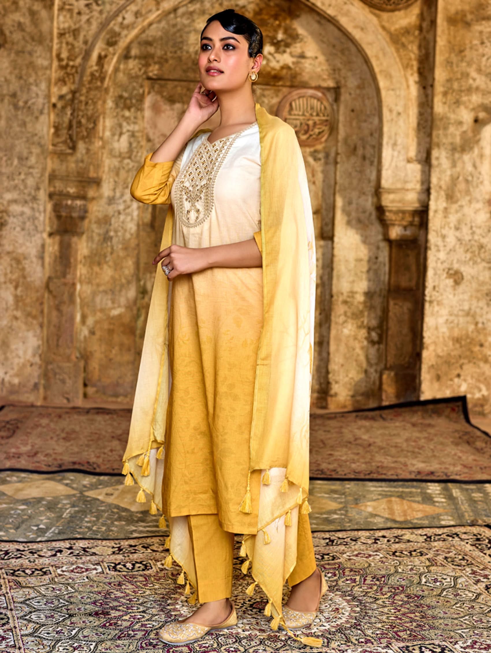 A woman wearing a yellow and beige digitally printed pure cotton suit set with a round neckline, three-fourth sleeves, side slits, and a dupatta.