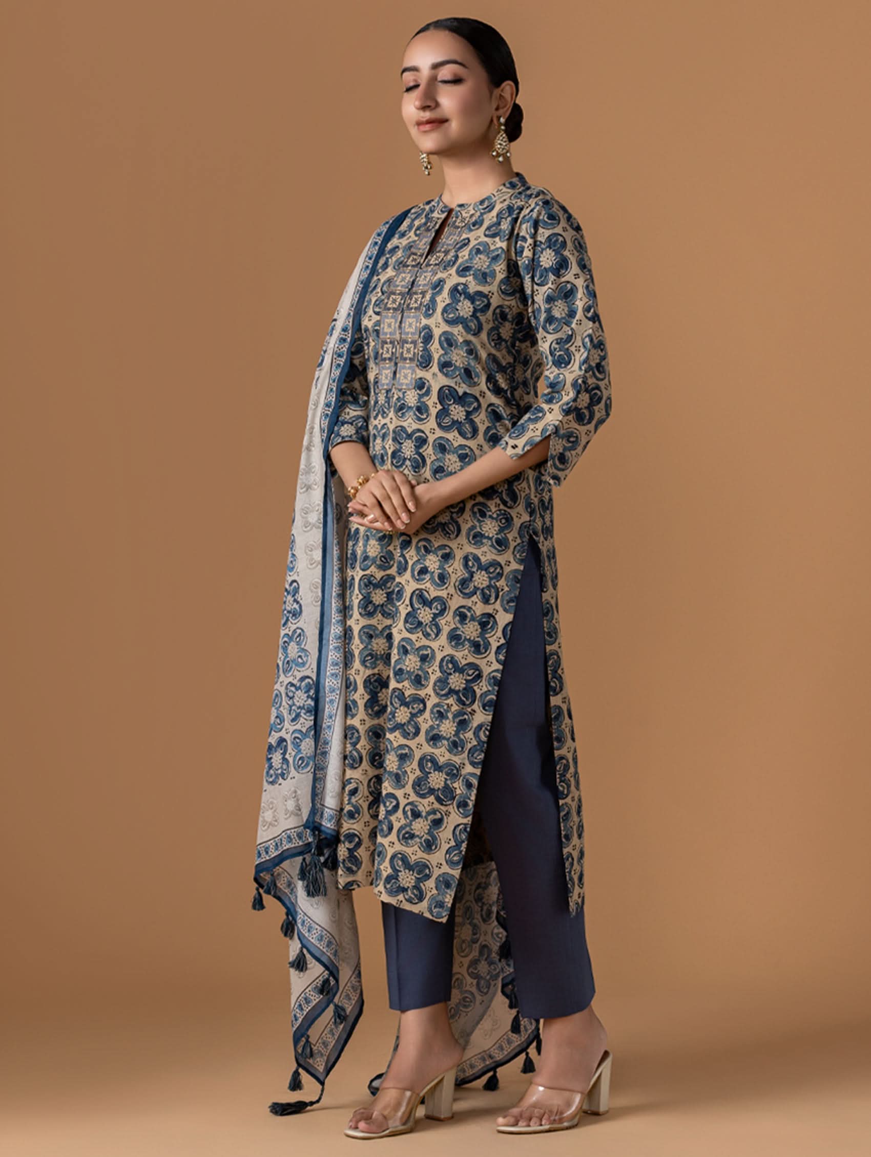 Woman wearing a blue and beige patterned outfit with a scarf against a brown background.