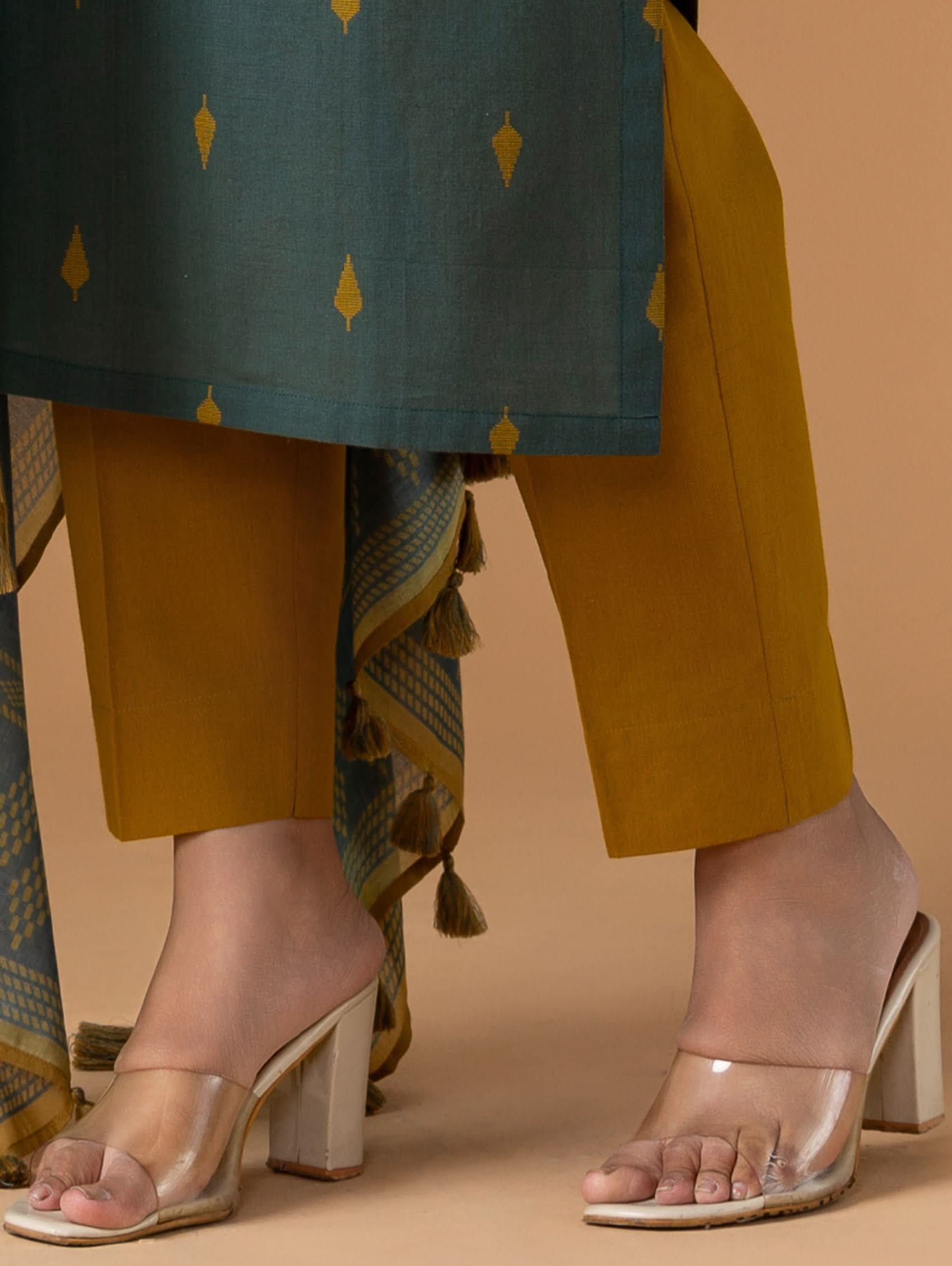 Person wearing mustard yellow pants and a green top with a pattern, paired with beige sandals on a beige background.