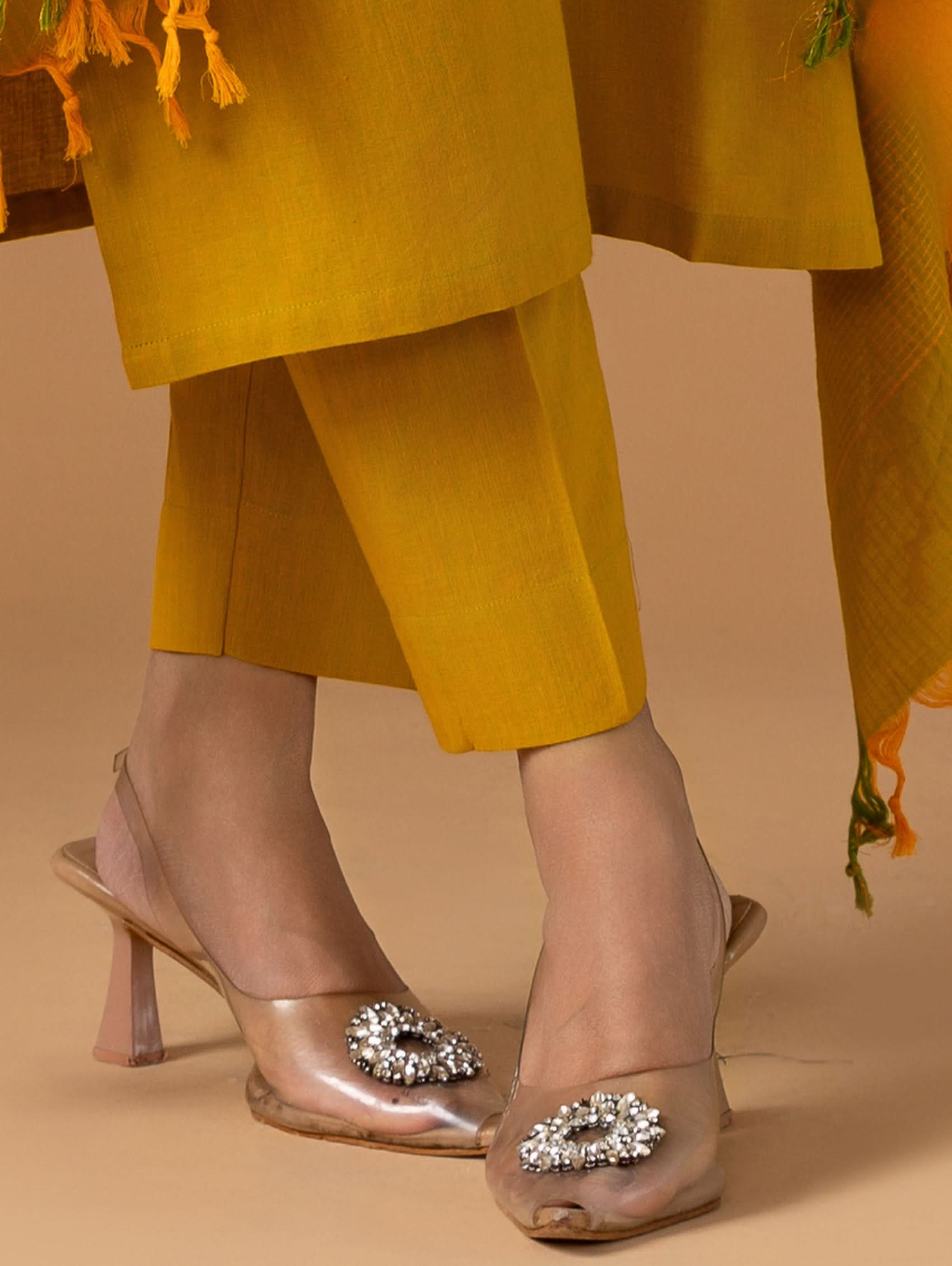 Clear high-heeled shoes with decorative elements worn with mustard yellow pants and a matching shawl.