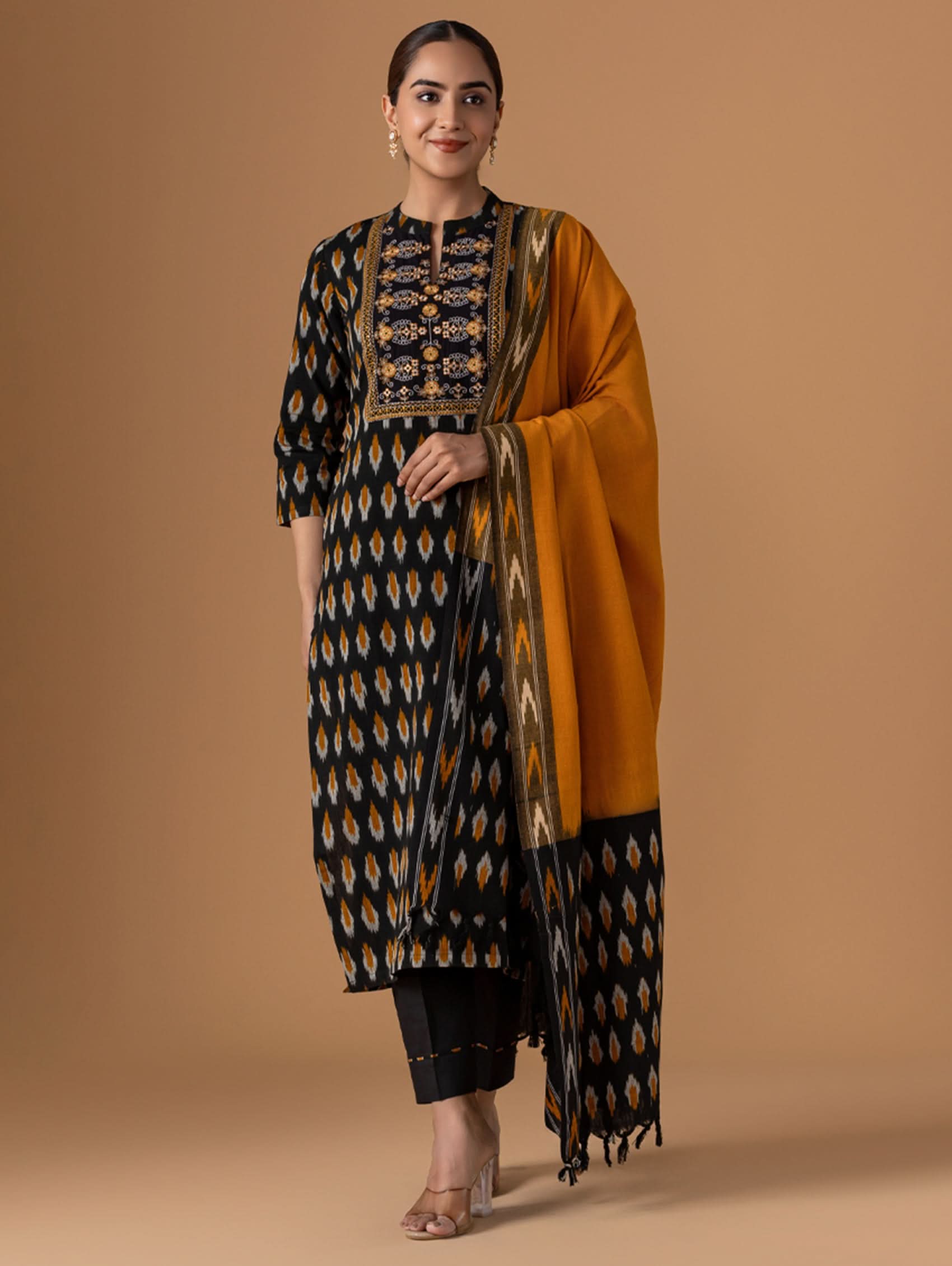 Woman wearing a traditional outfit with a black and gold patterned kurta and mustard yellow dupatta on a beige background