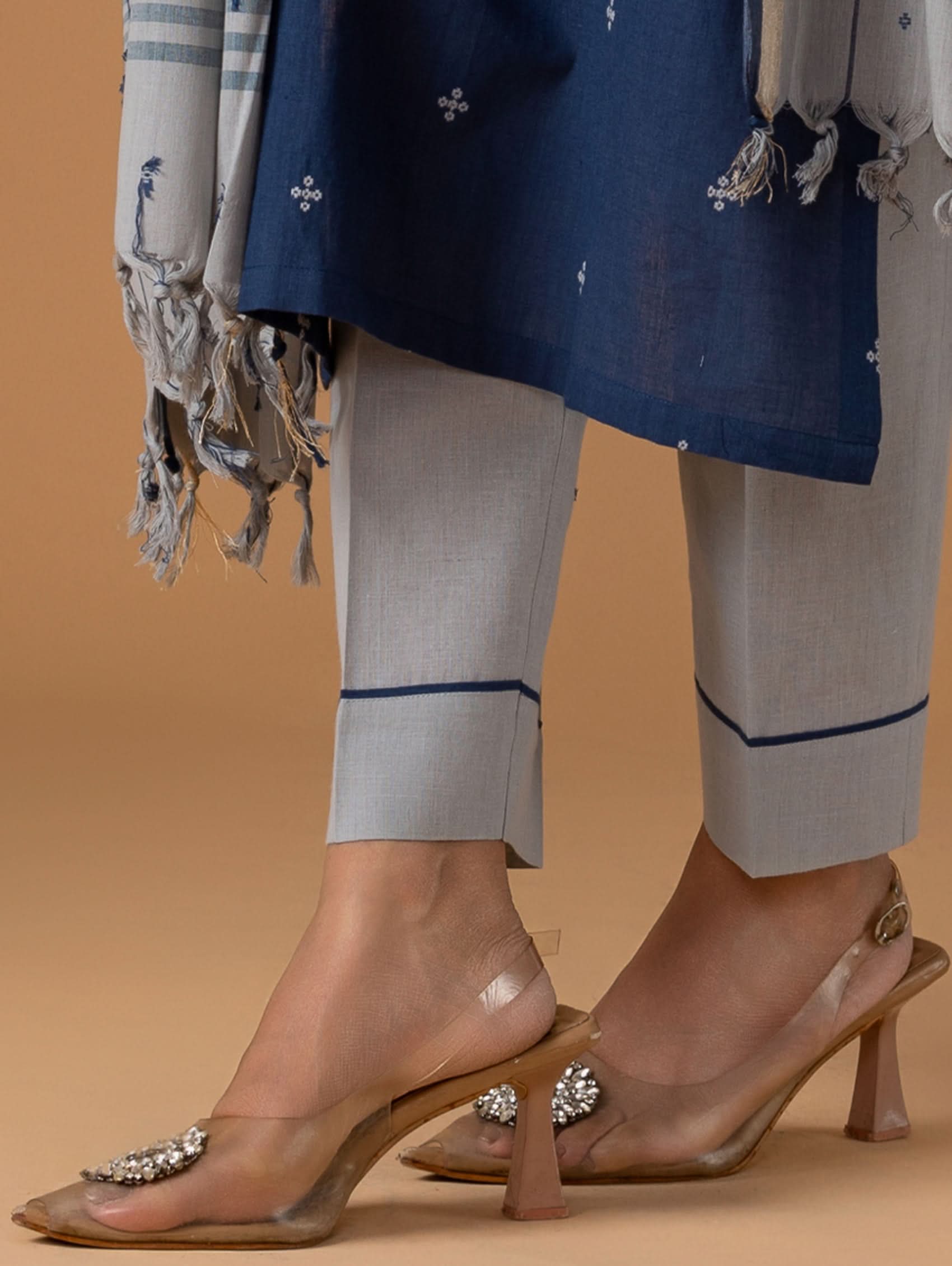 Person wearing a blue top, gray pants, and clear high-heeled shoes with embellishments on a beige background