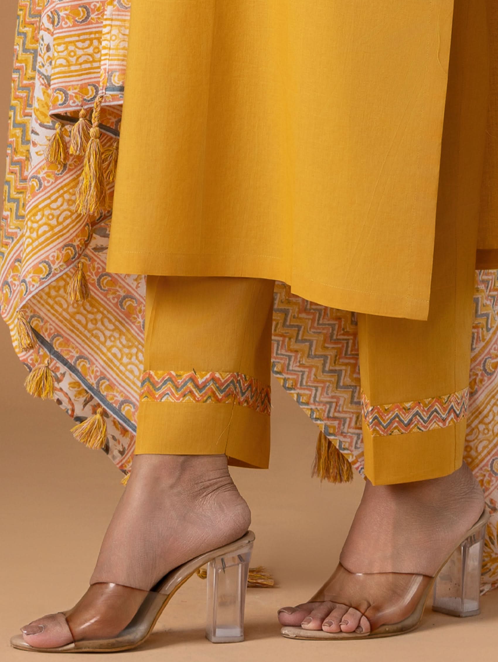 Person wearing mustard yellow pants with patterned trim and transparent high-heeled shoes on a beige background