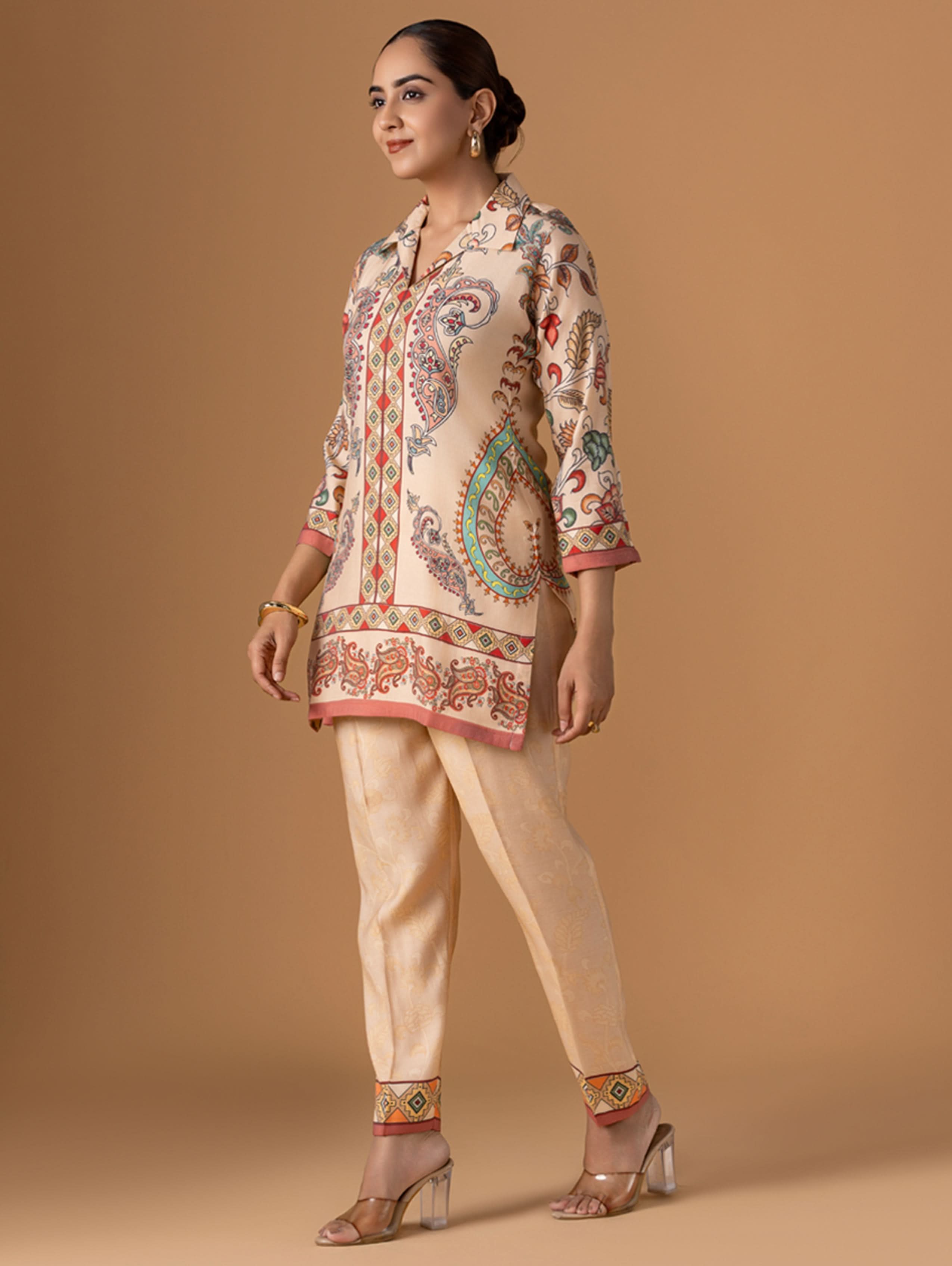 Woman wearing a traditional outfit with a patterned top and beige pants on a brown background