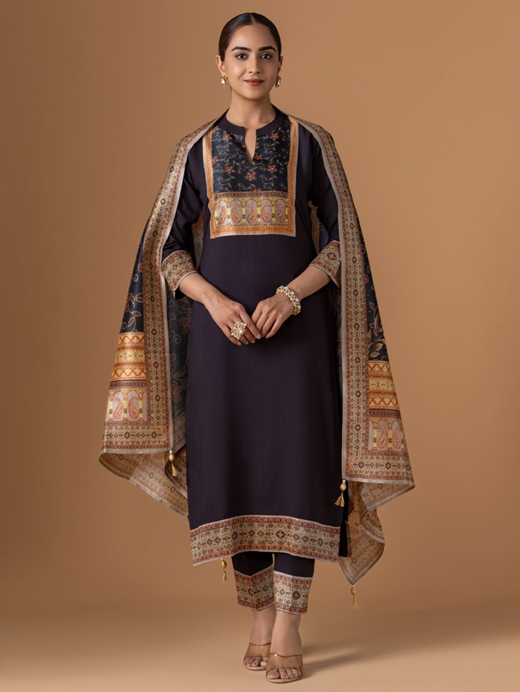 Woman wearing a traditional outfit with a dark blue top and pants, and an embroidered shawl on a brown background.