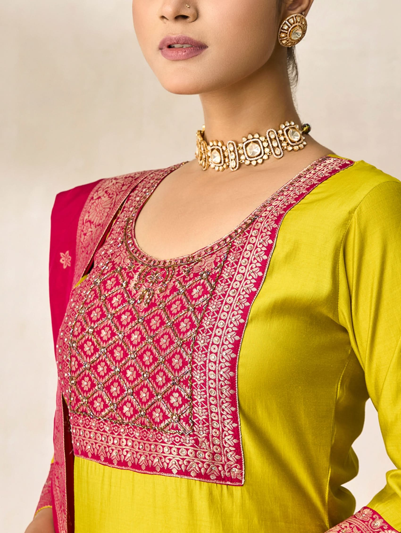 Woman wearing a yellow outfit with pink embroidered blouse and gold necklace against a neutral background