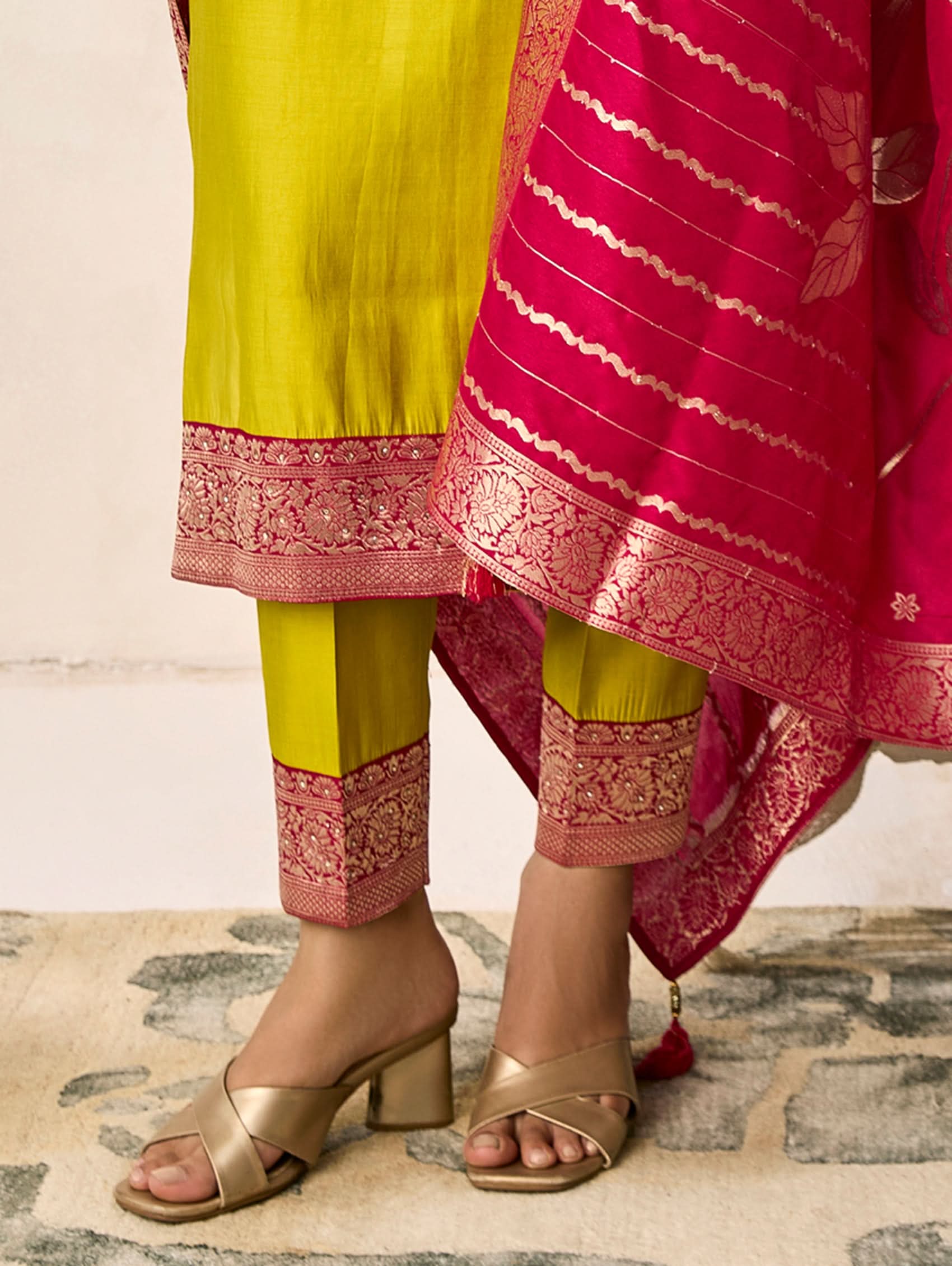 Person wearing a bright yellow outfit with pink embroidery and gold sandals on a patterned rug.