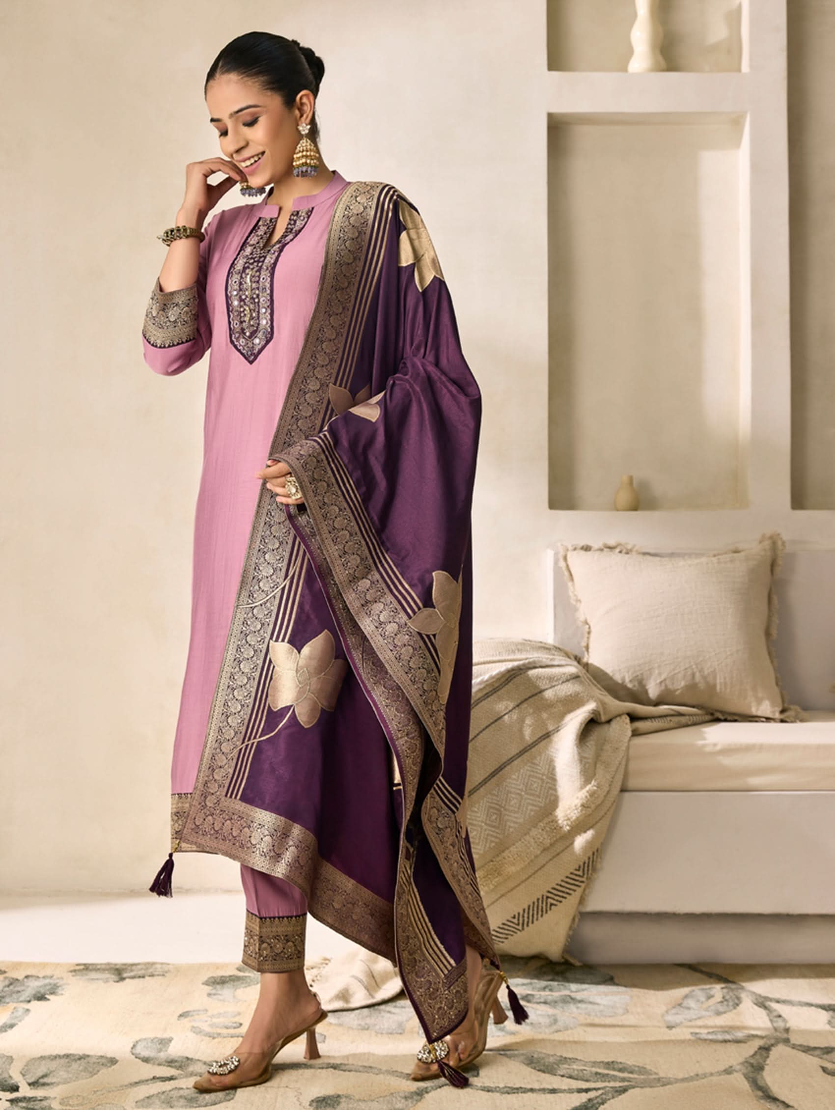 Woman wearing a pink and purple traditional outfit with a matching dupatta in a living room setting.