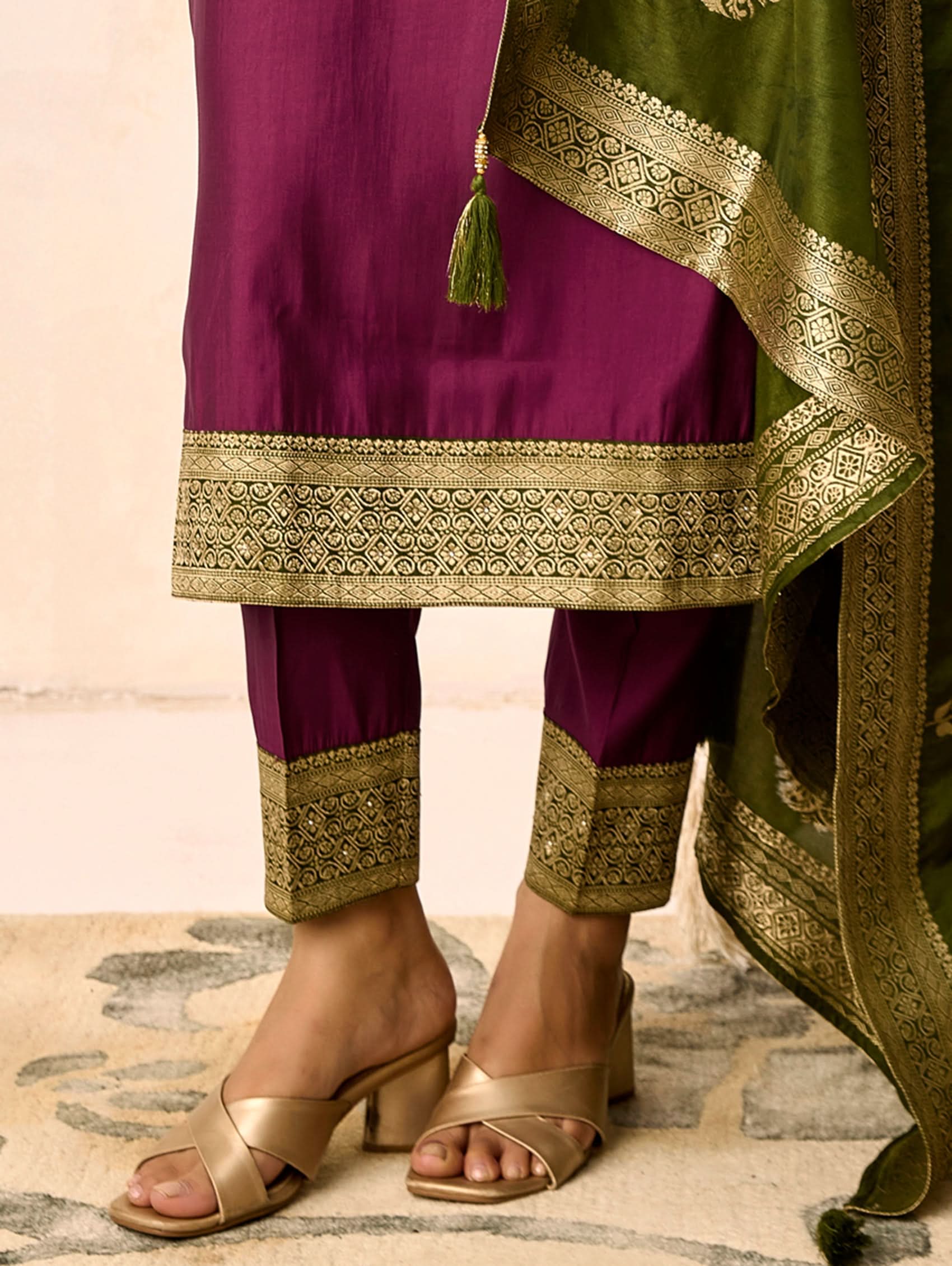 Traditional outfit with purple and green fabric, gold embroidery, and sandals on a neutral background