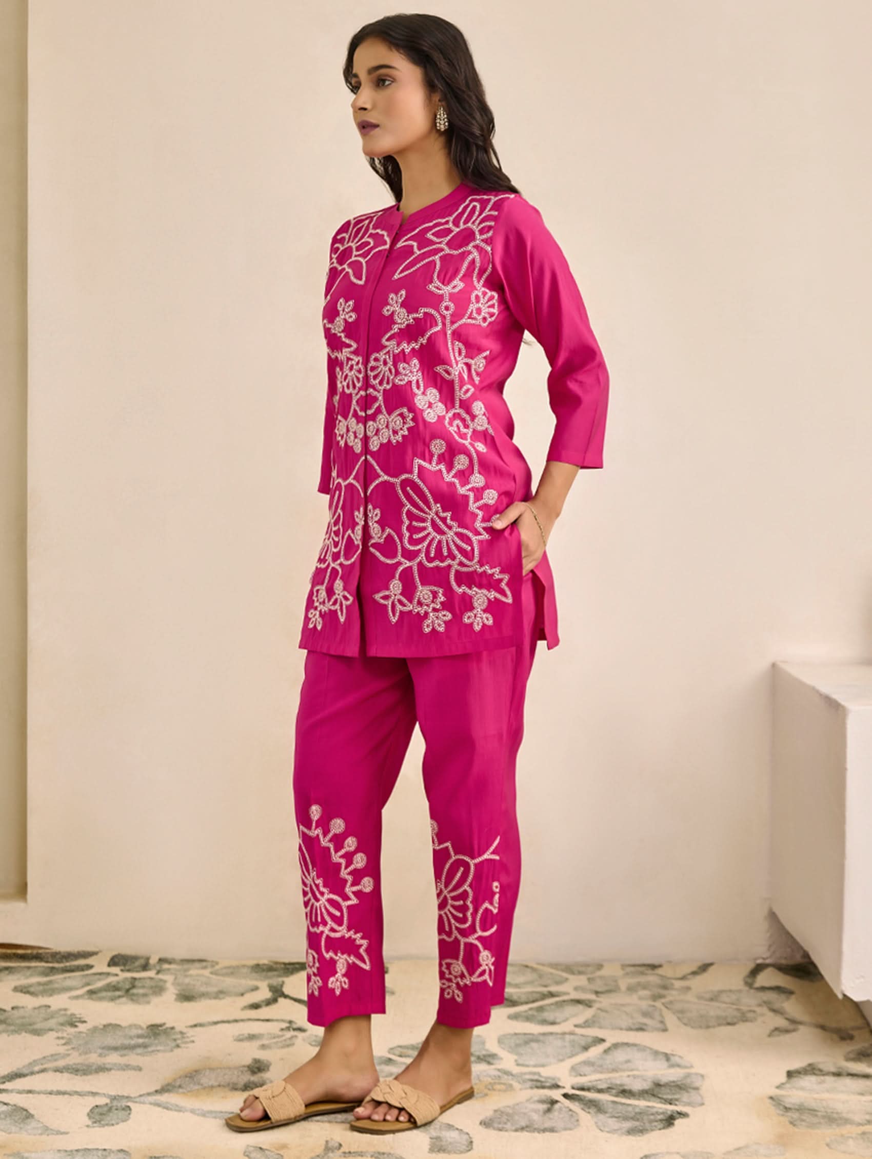 Woman wearing a pink embroidered outfit in a room with a patterned rug.