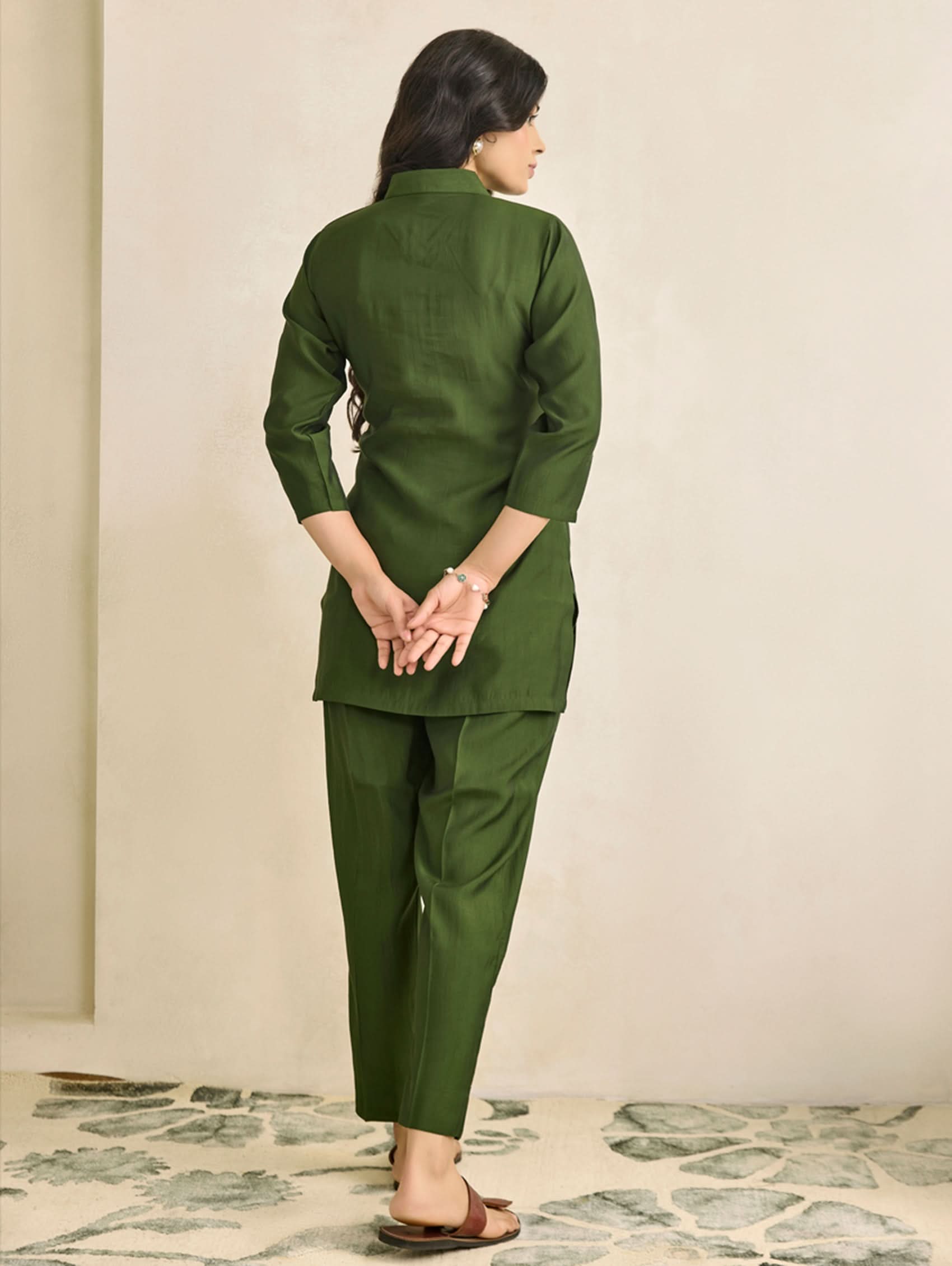 Person wearing a green traditional outfit standing against a beige wall.