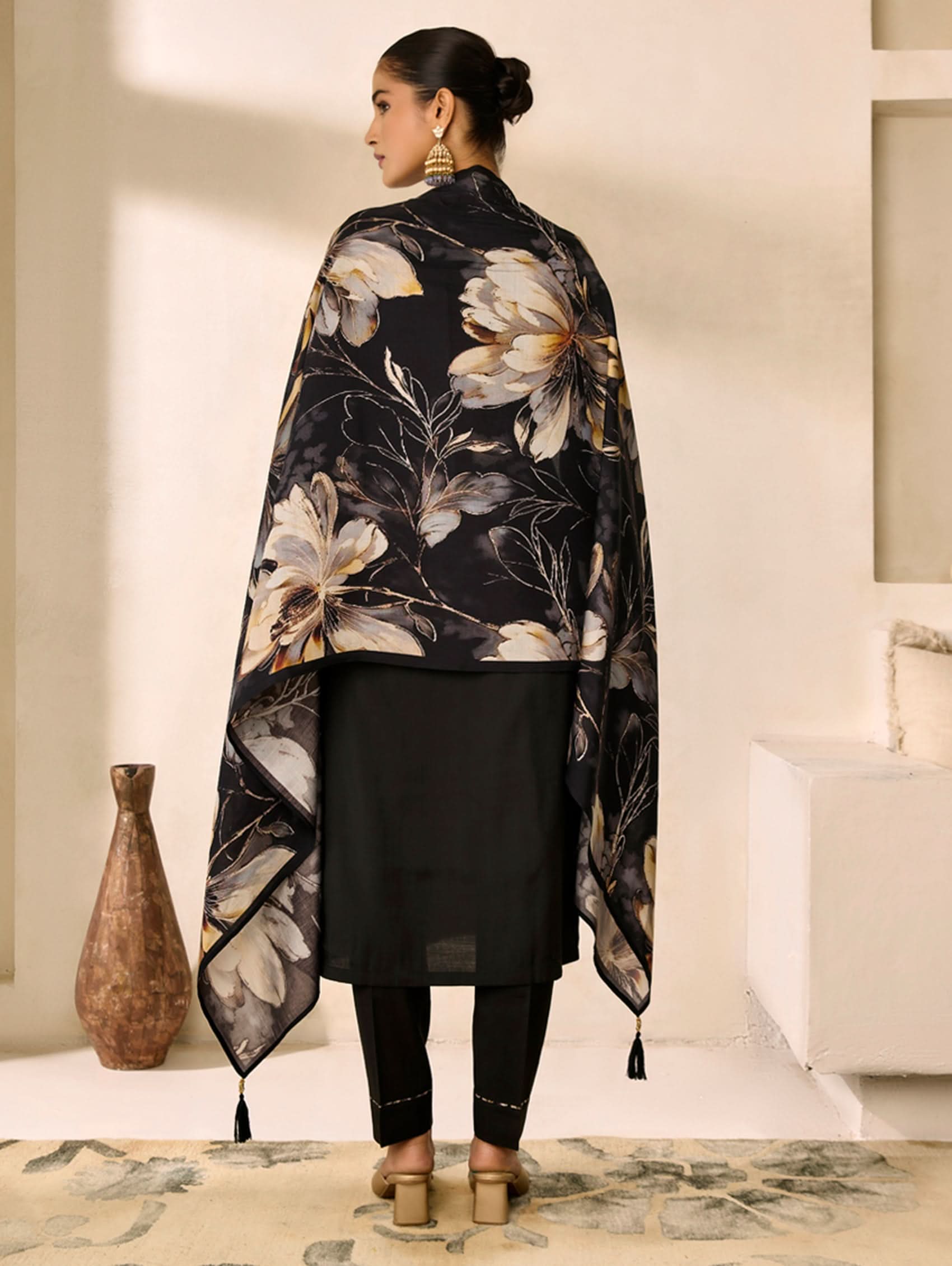 Woman wearing a black floral shawl in a room with a vase and rug.