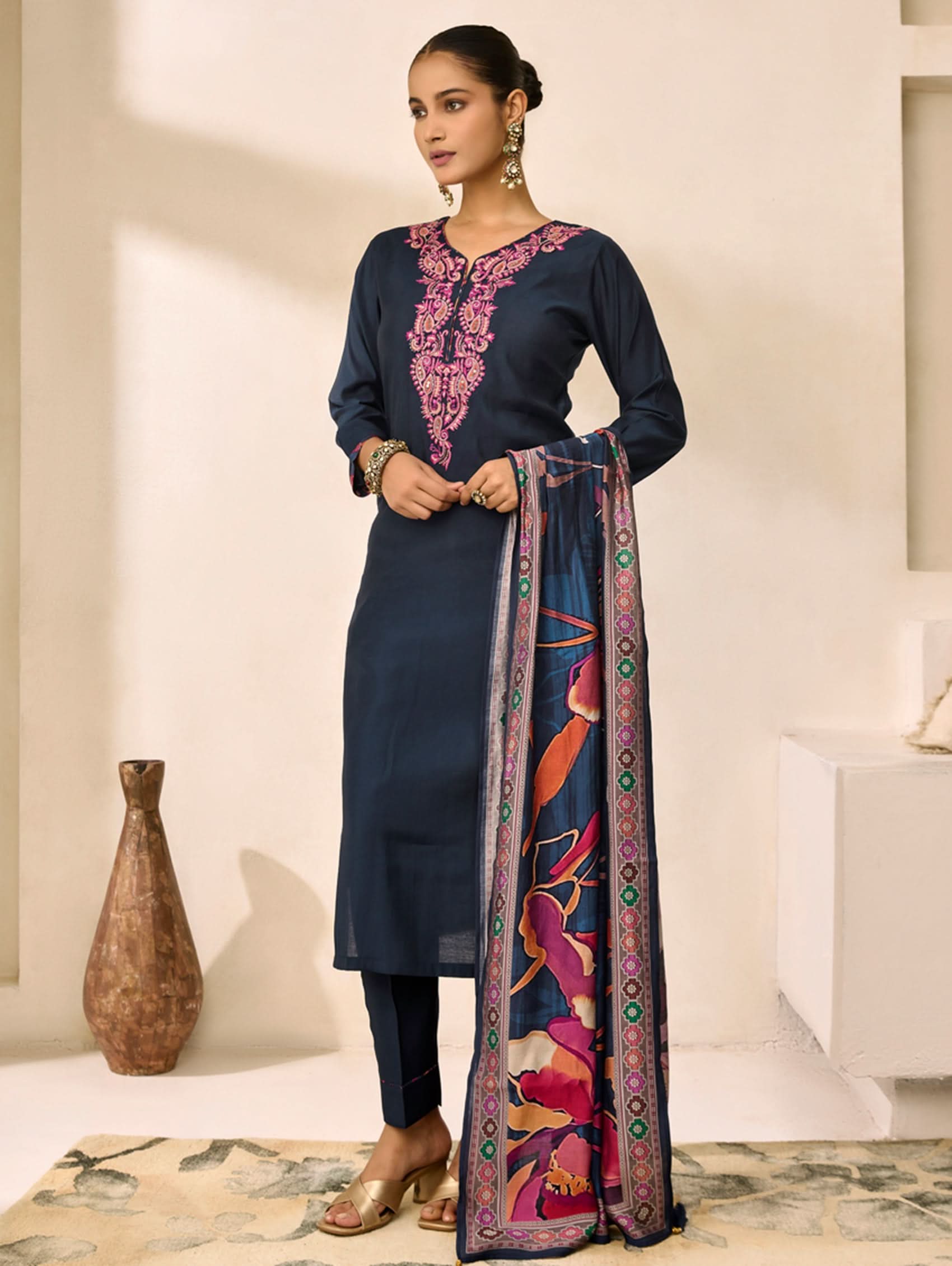 Woman wearing a navy blue kurta with pink embroidery and a colorful dupatta in a minimalistic setting.