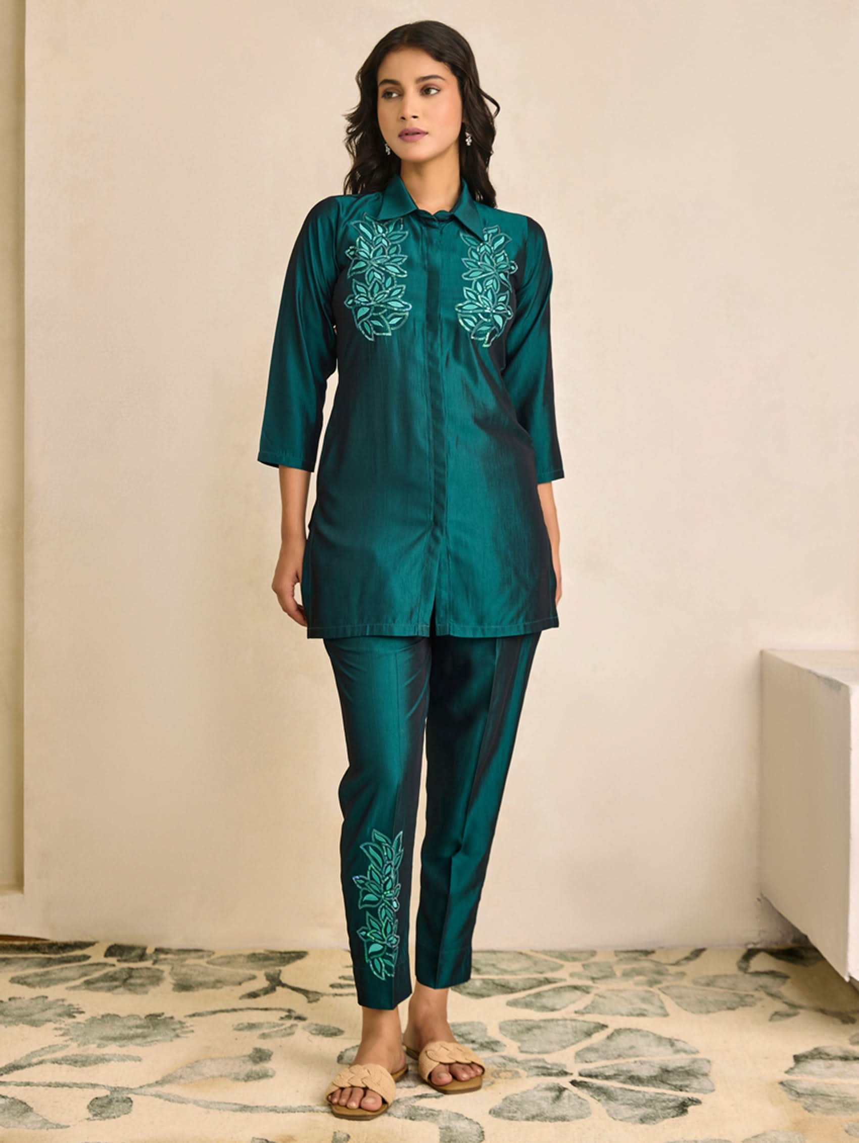 Woman wearing a teal outfit with embroidery in a room with a patterned rug.