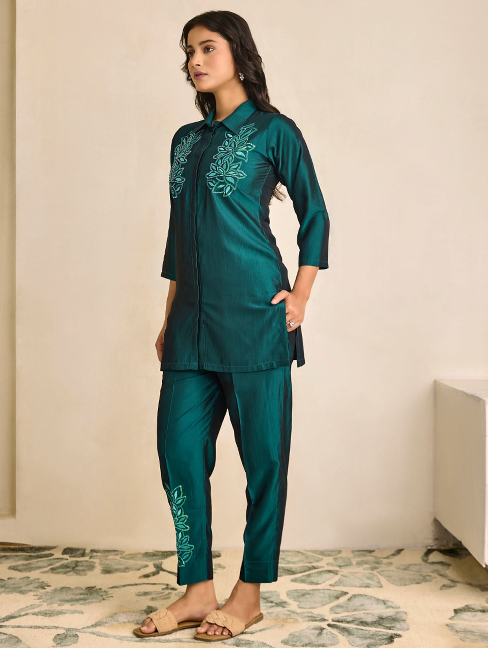 Woman wearing a teal embroidered outfit standing in a room with a neutral background.