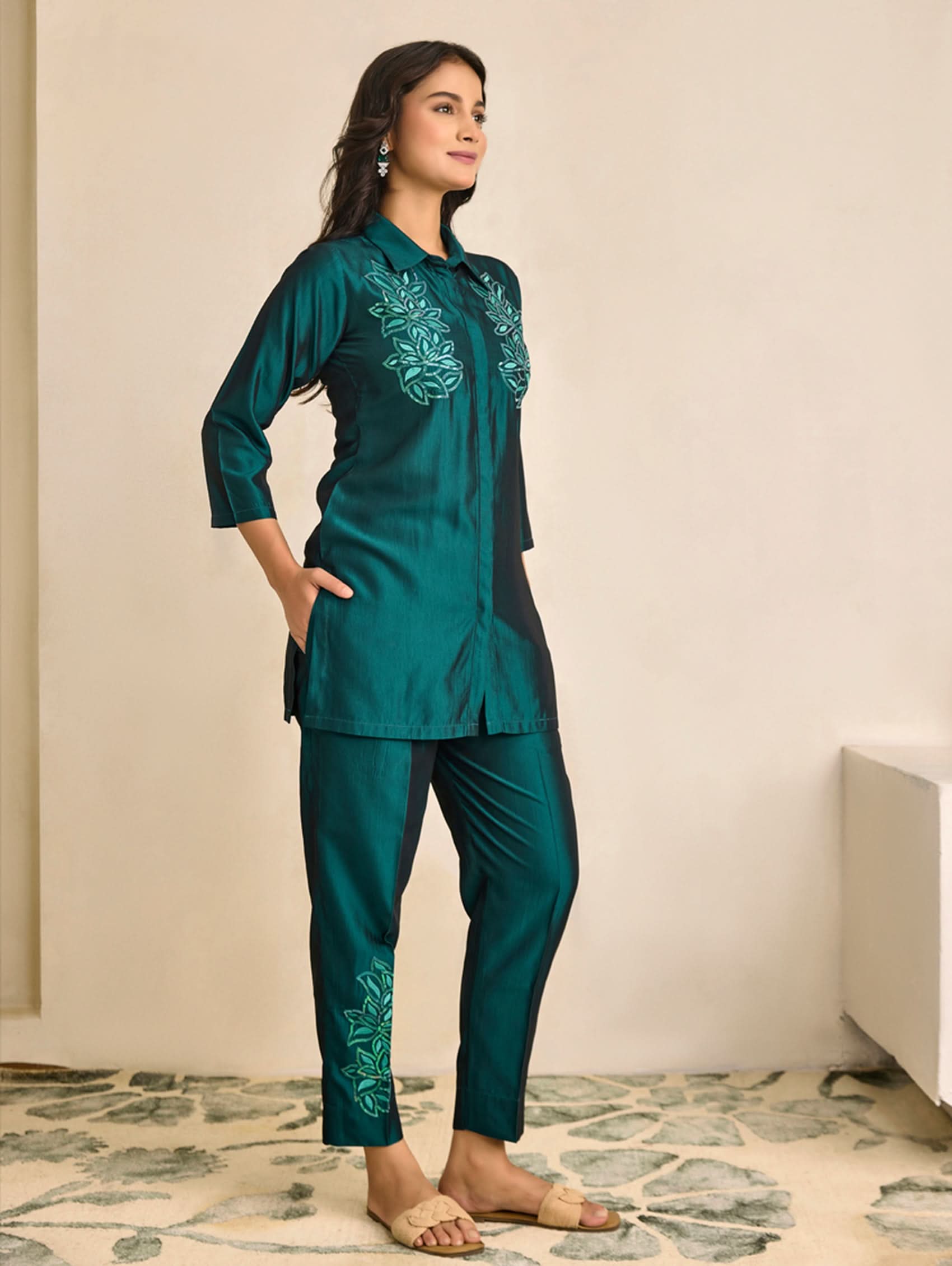 Woman wearing a teal embroidered outfit standing against a beige wall.