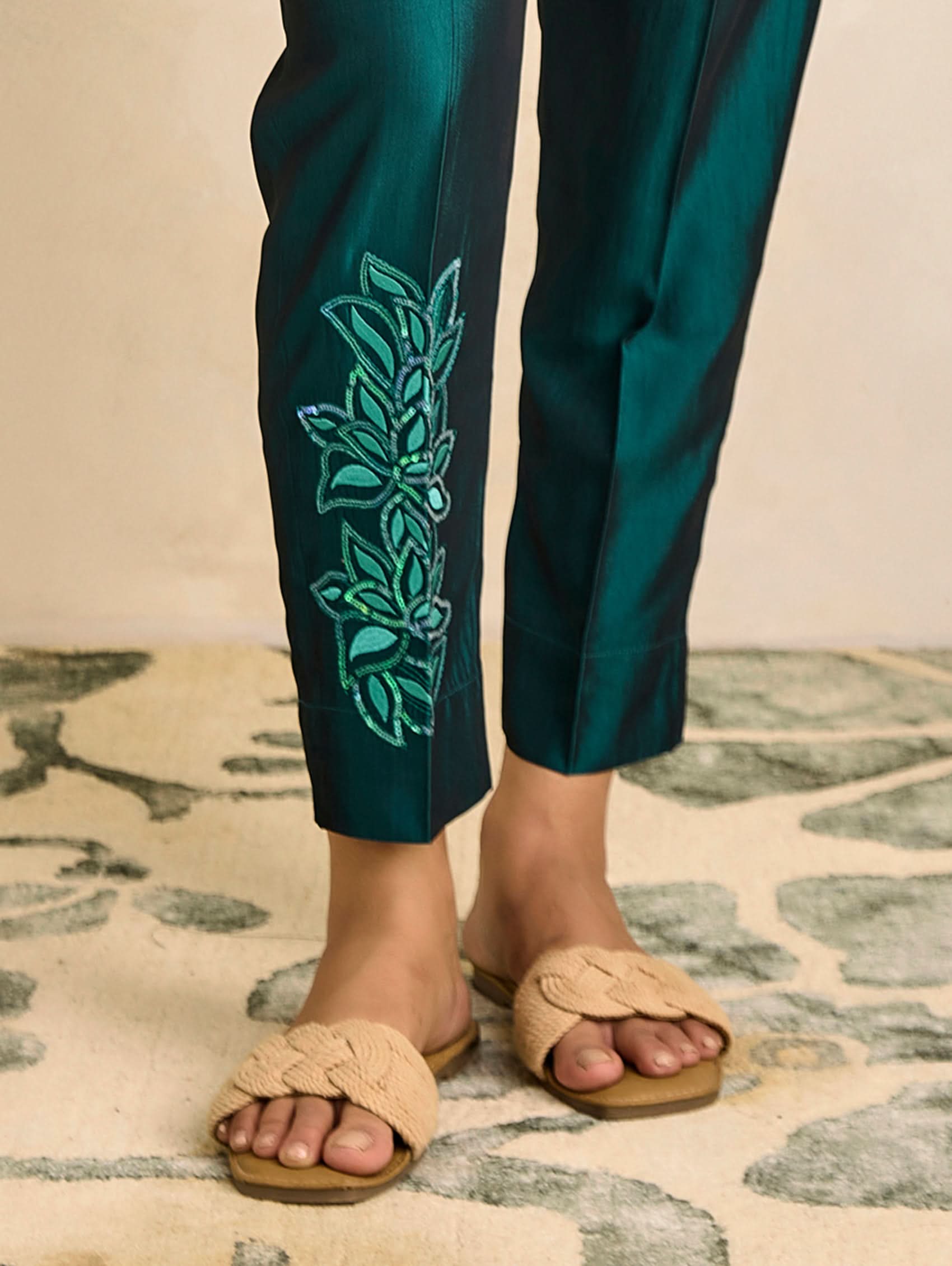 Teal pants with green embroidery and beige sandals on a patterned rug